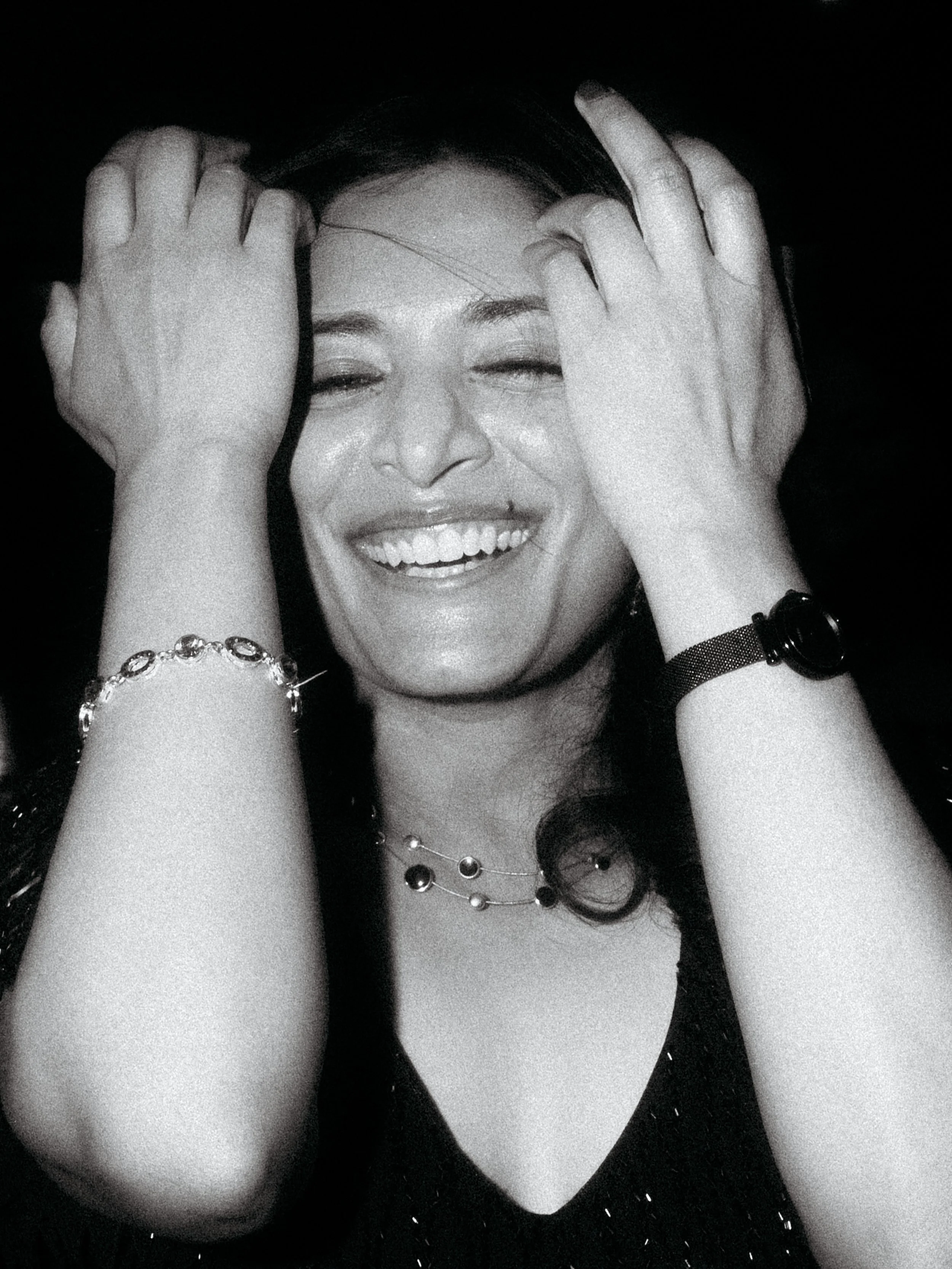 Black and white photo of a woman with a big smile, eyes closed, hands on her head, wearing jewelry including necklaces, bracelets, and a watch.