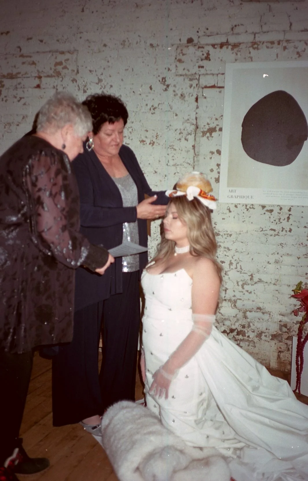 A woman in a white wedding dress sitting on the floor with a large skirt, wearing a hat with orange and white decorations, during a wedding ceremony with two women standing in front of her.