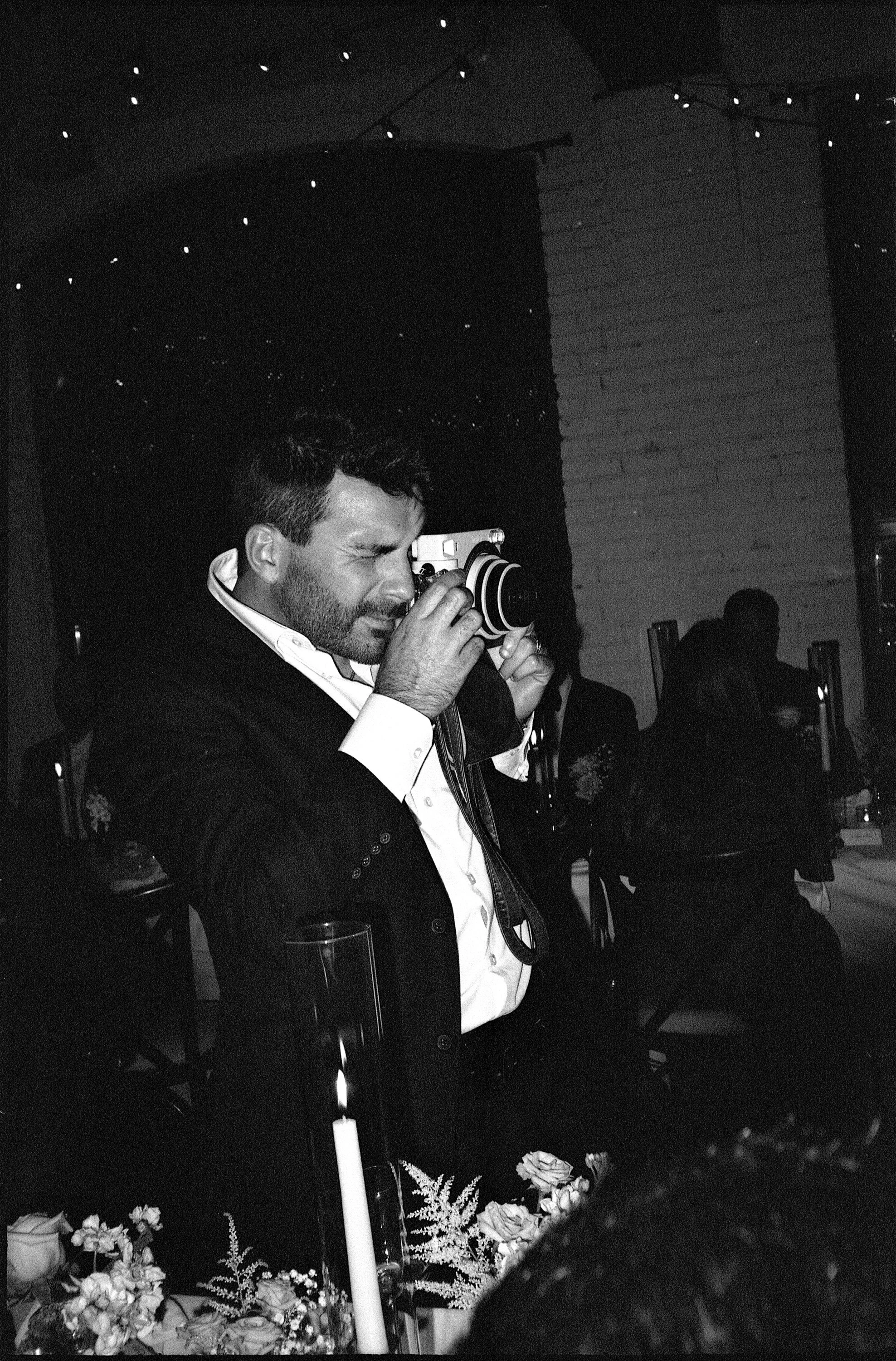 A man with dark hair and a beard, dressed in a white shirt and dark jacket, taking a photo with a vintage camera at a social event decorated with flowers and candles, with several other people seated in the background.