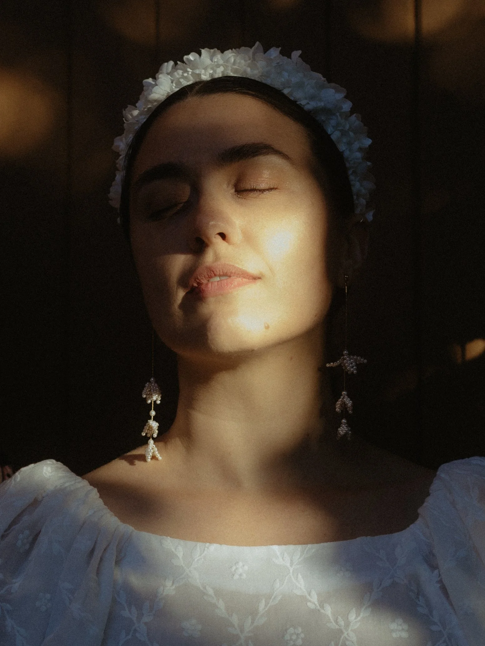 A woman with closed eyes wearing a white flower crown and white embroidered dress, with long earrings, standing in a dimly lit environment.