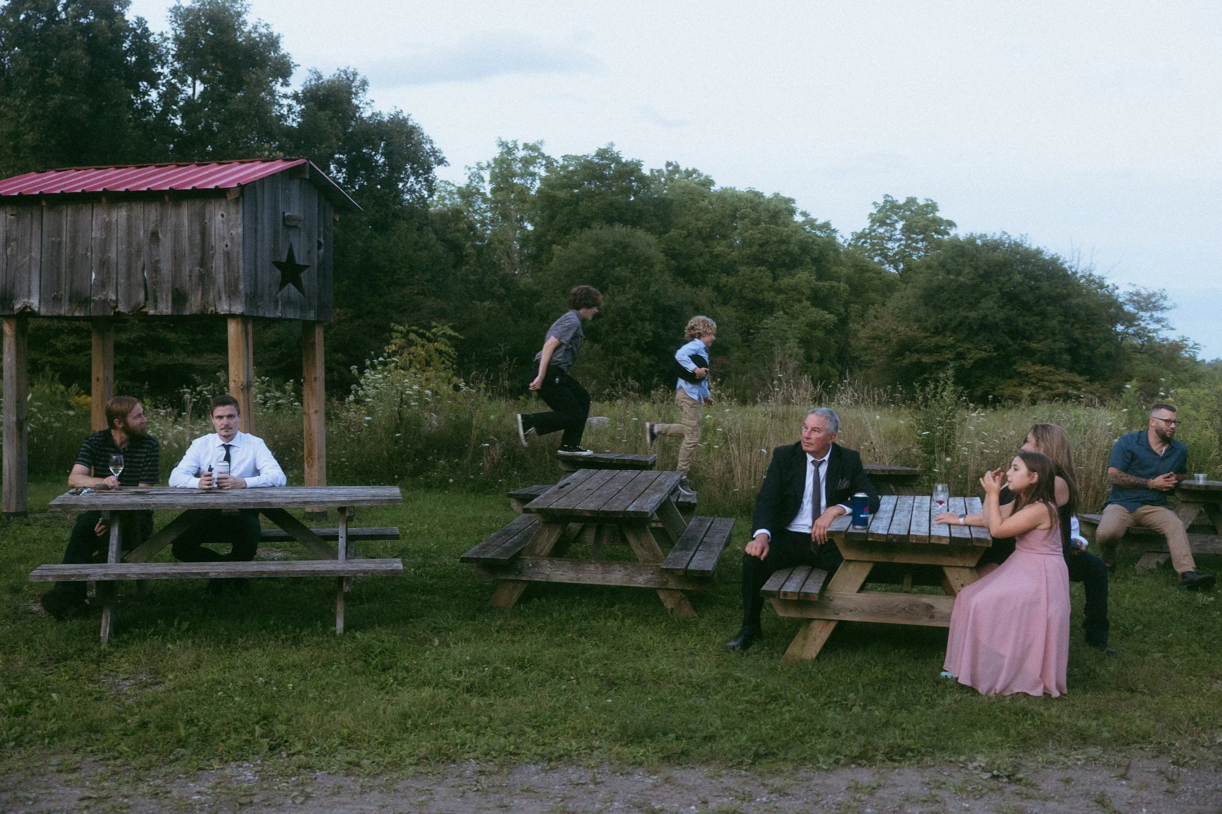People sitting at outdoor picnic tables, with two children jumping between the tables, in a grassy area near a small wooden shed with a star, surrounded by trees.