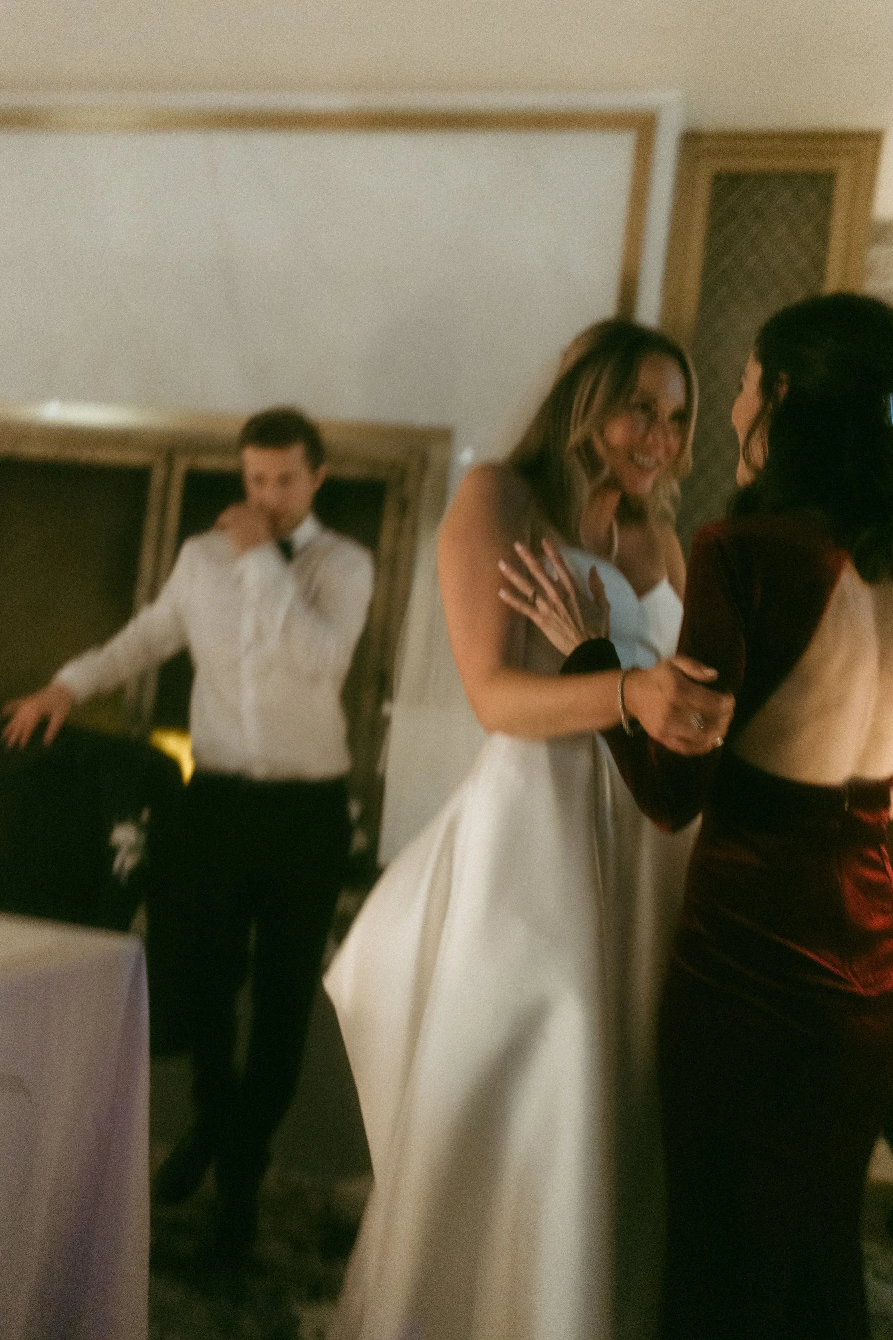 A bride in a white wedding gown interacts with another woman at a wedding reception, while a man in a white shirt stands in the background.