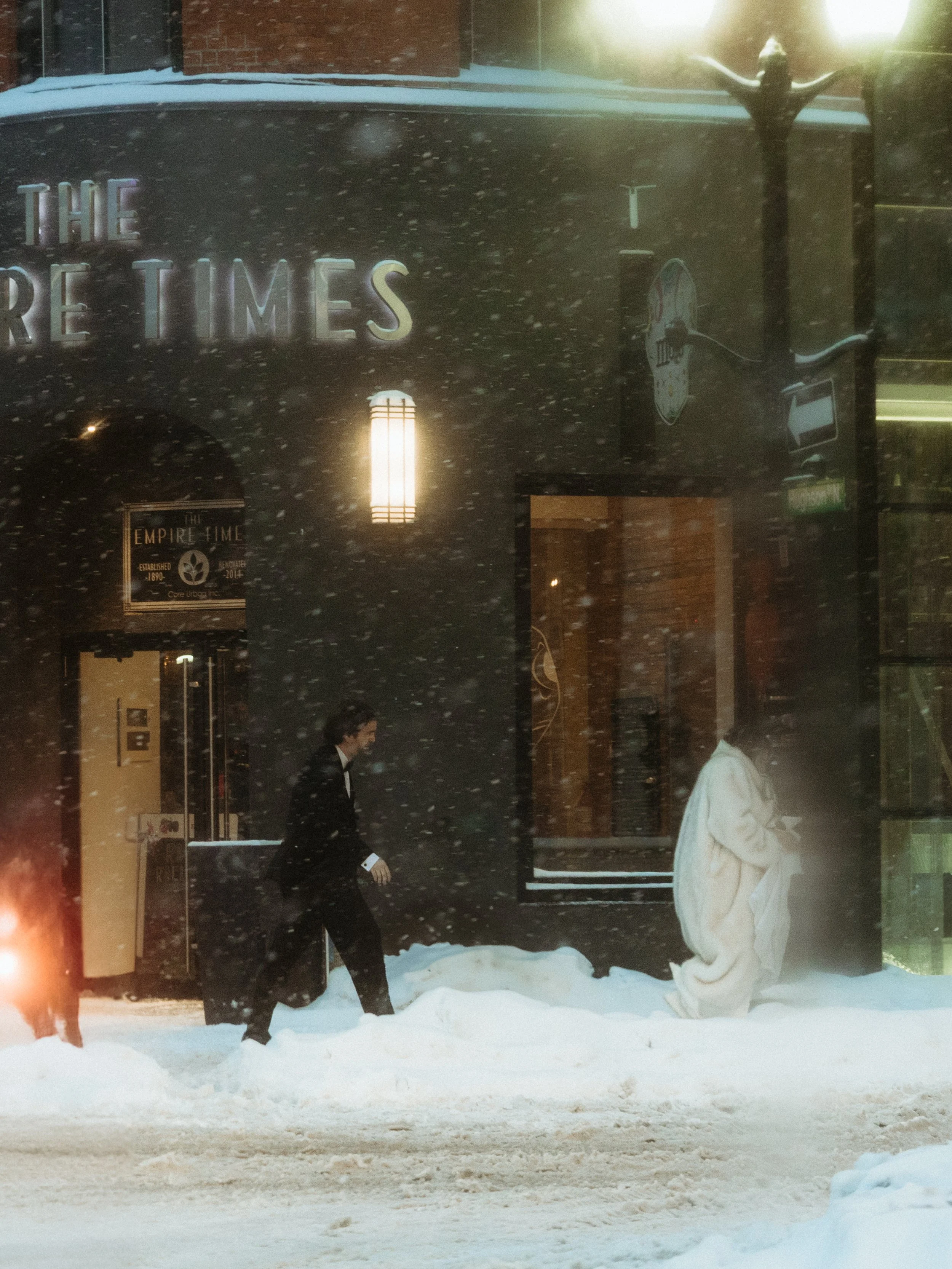 People dressed in winter clothing walking through snow in front of a building with a sign that says 'The New York Times' during snowfall at dusk or night.