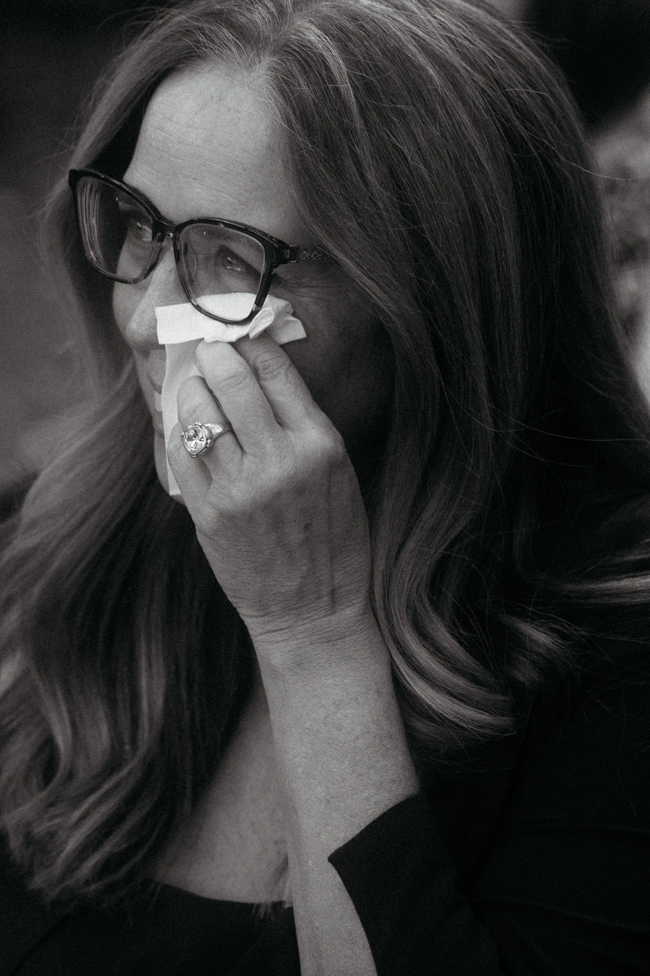 A woman with long hair and glasses is wiping away tears with a tissue.