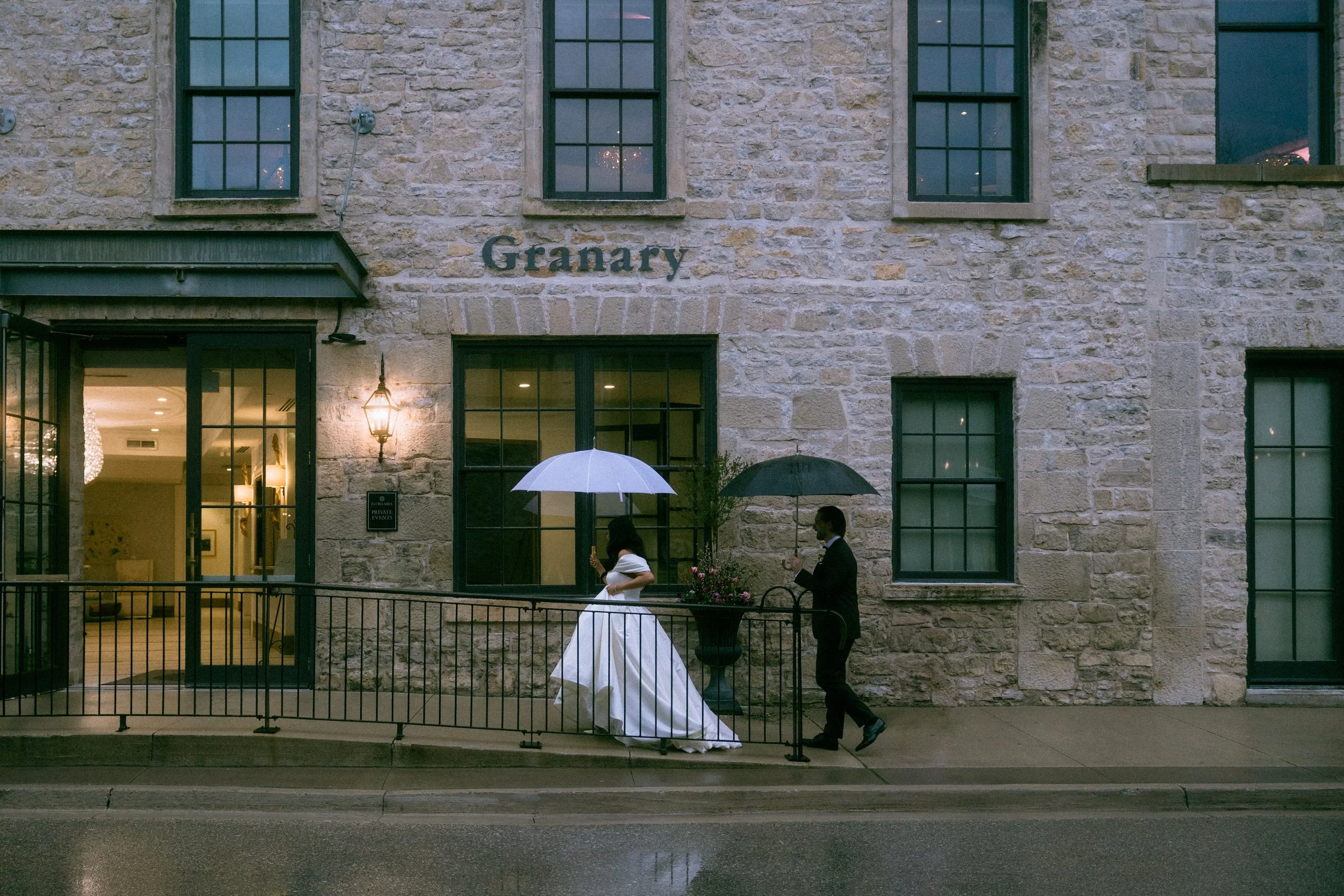 A woman in a wedding dress and a man in a suit hold umbrellas while walking on a sidewalk in front of a stone building with large windows and a sign that says "Granary." The woman is looking at her phone, and the man is looking at something in his ha
