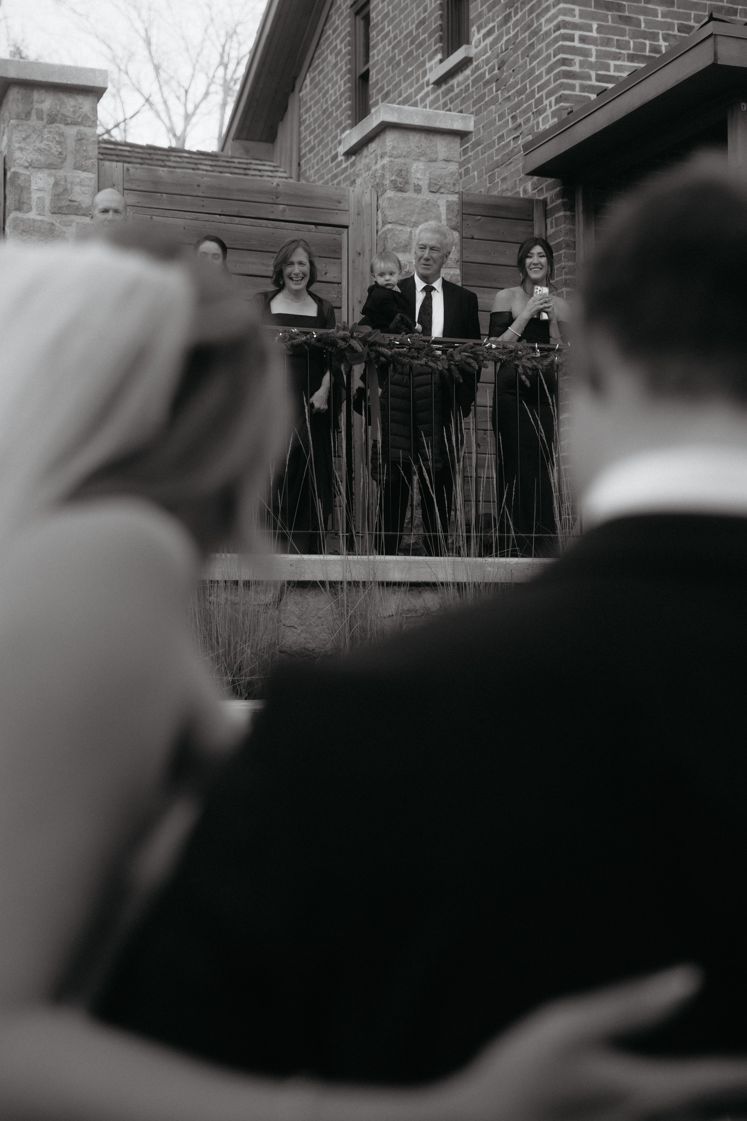 People gathered on a balcony watching an outdoor event, with some individuals smiling and holding phones, including a man in a suit and a woman with a child, against a brick house and wooden fence background.