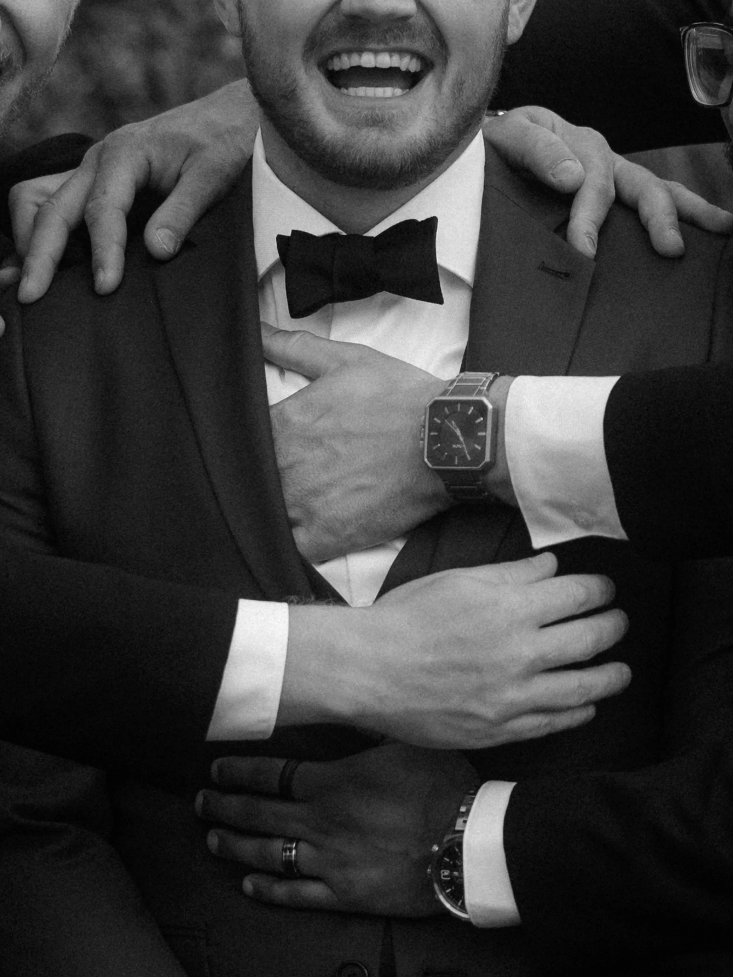 A group of men in tuxedos, embracing each other, with one man smiling widely. Hands are placed on each other's shoulders and chests, and watches are visible on their wrists.