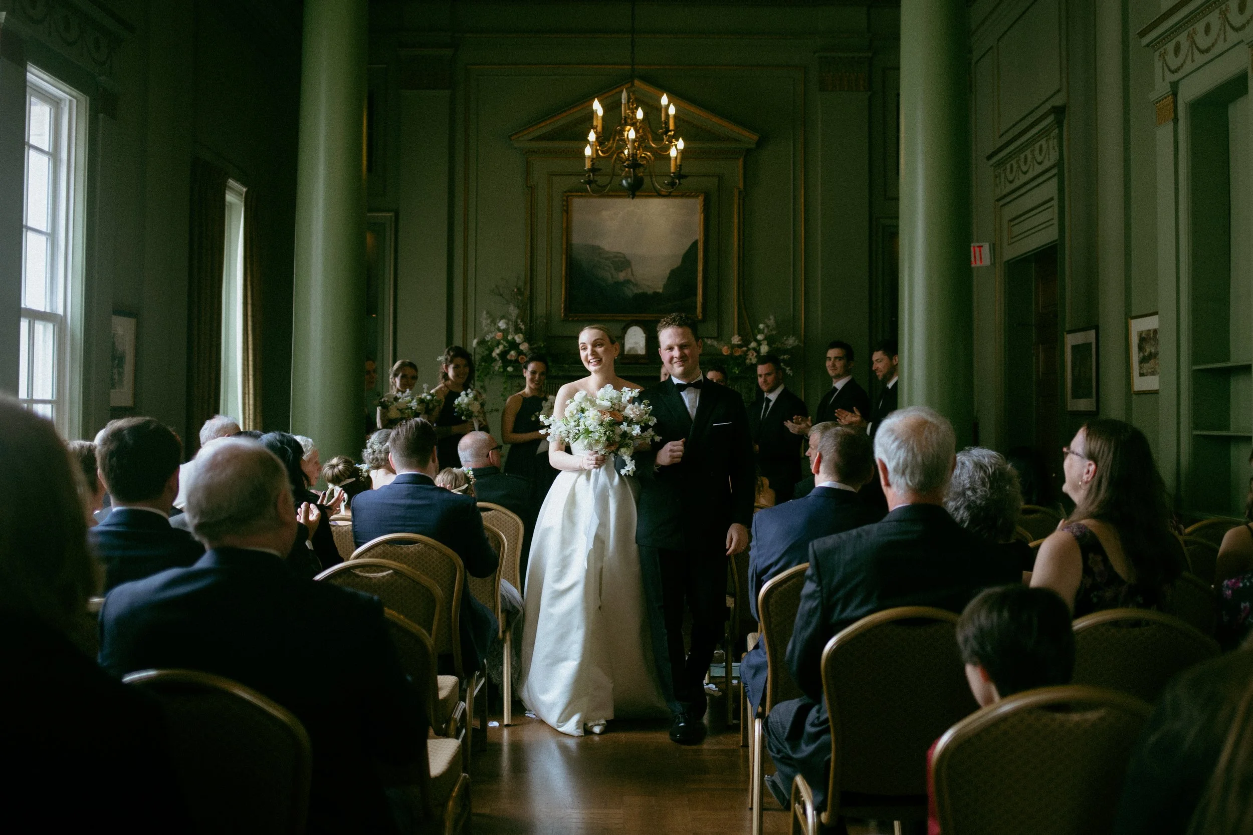 A bride in a white wedding dress holding a bouquet and a groom in a black tuxedo walking down the aisle during a wedding ceremony in a decorated room with green walls and a chandelier, surrounded by seated guests and a bridal party standing in the background.