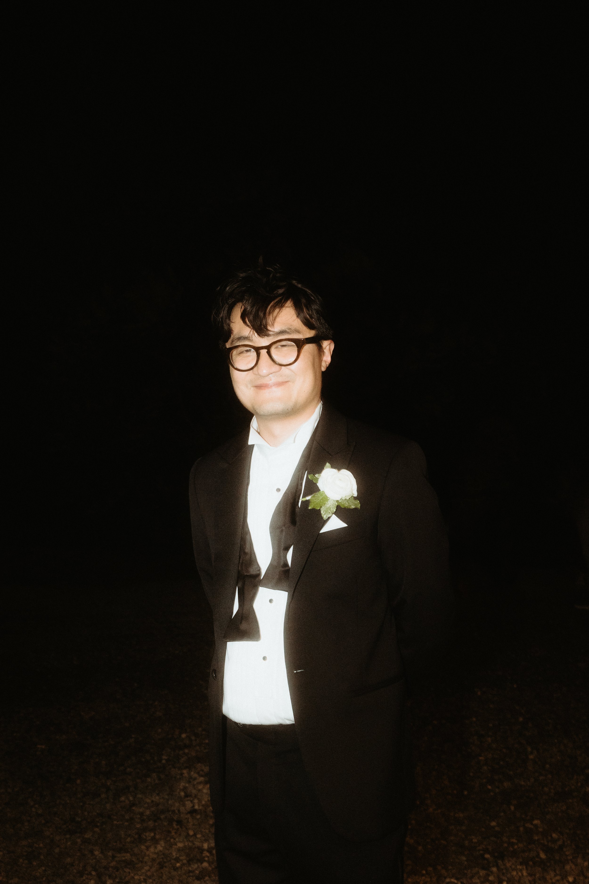 A man wearing glasses in a tuxedo with a white boutonniere, smiling at night against a dark background.