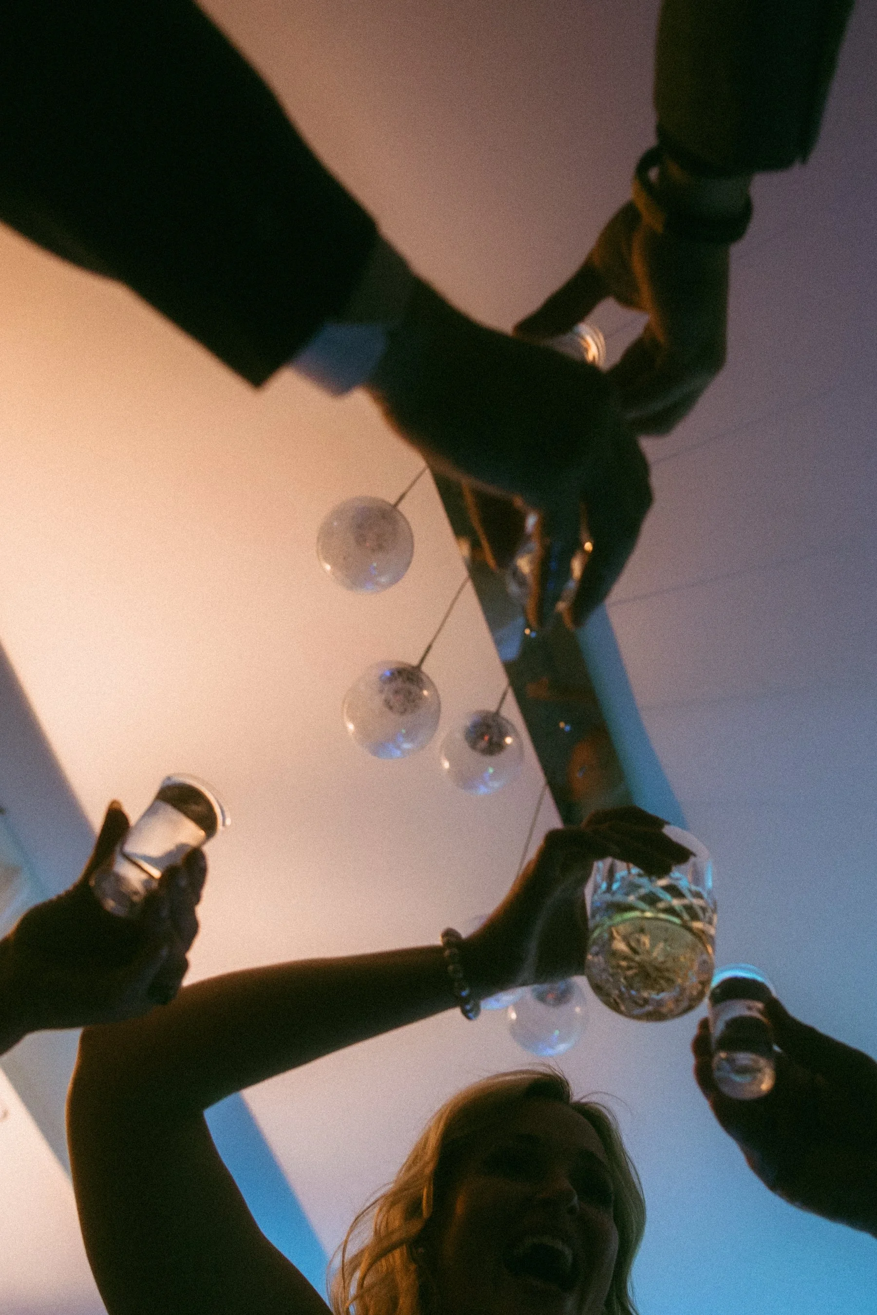 People celebrating, raising glasses in a toast indoors with hanging spherical light fixtures and a smiling woman in the foreground.