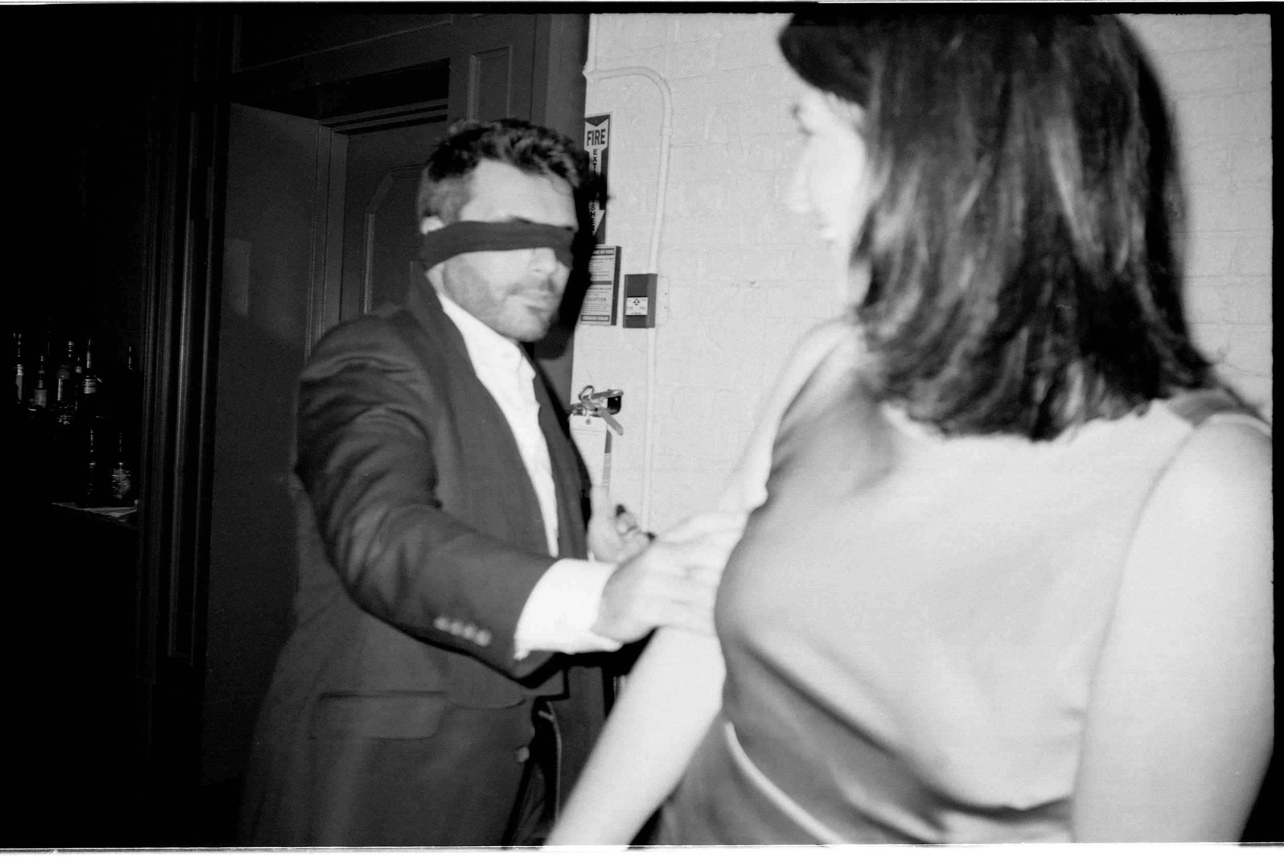 A black-and-white photo of a man in a suit and a woman in a top, holding hands. The man is blindfolded and appears to be reaching out to the woman.