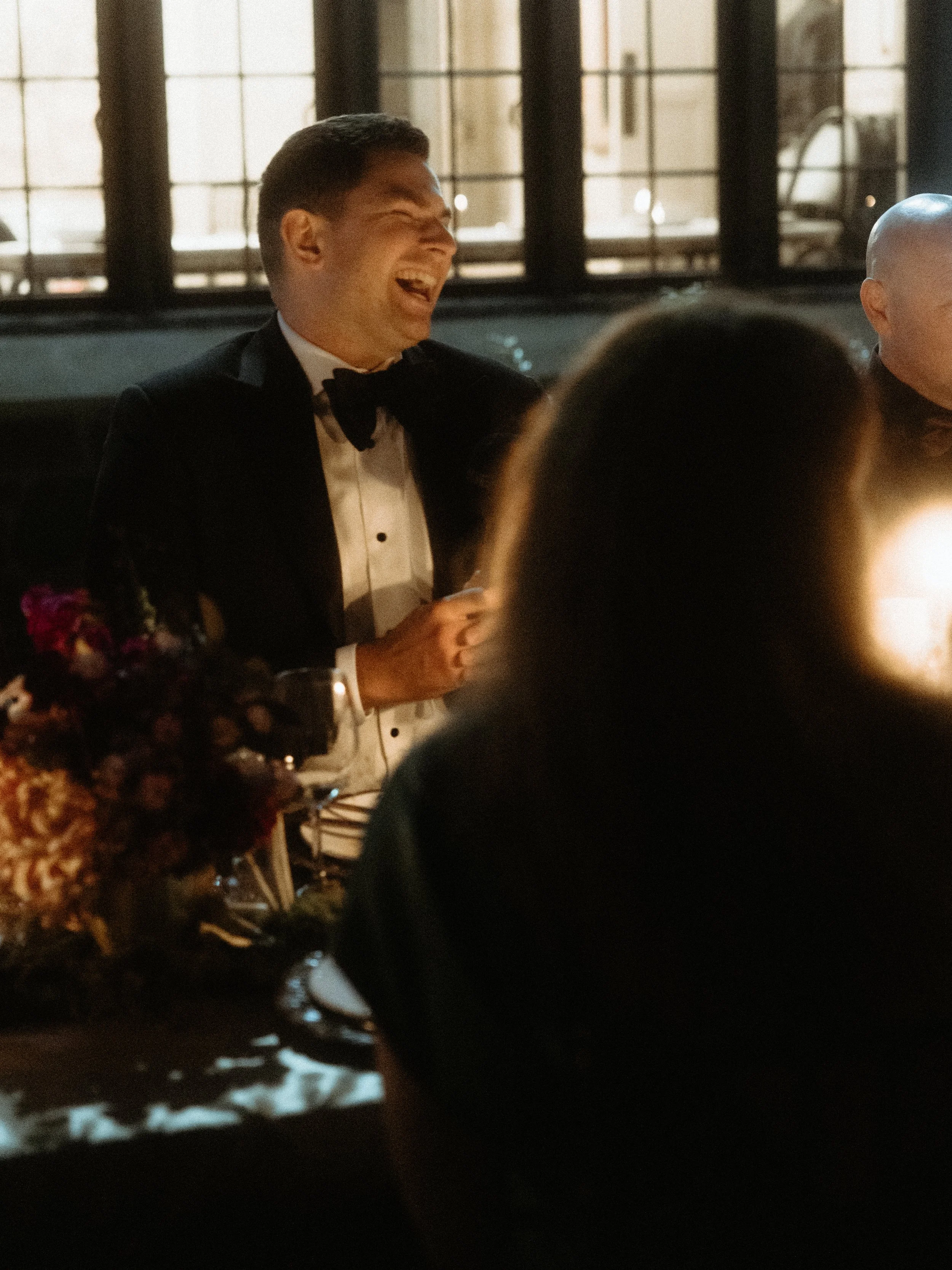 A man in a tuxedo laughing at a formal event, surrounded by other guests and dim lighting.