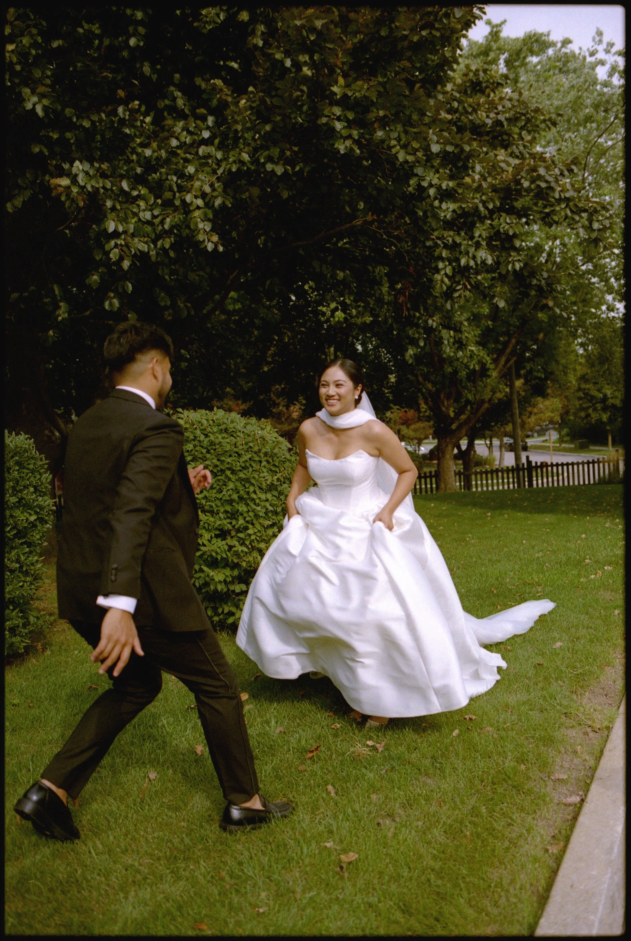 A bride in a white wedding dress Playfully lifts her dress as a groom in a black suit approaches her, set outdoors in a grassy area with trees and a sidewalk in the background.