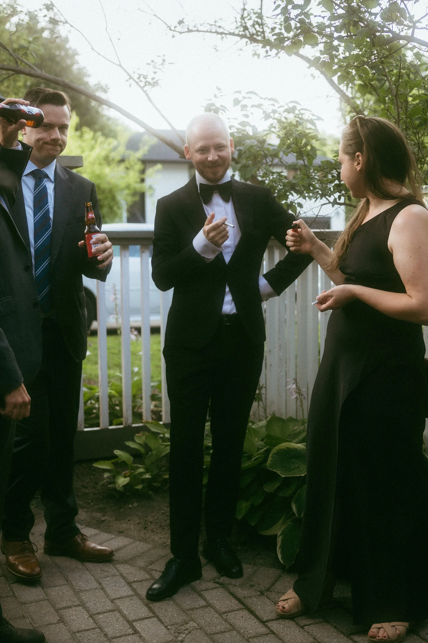 Group of people at a social gathering, with men in suits and women in an elegant dress, outdoors near a white fence and green plants.