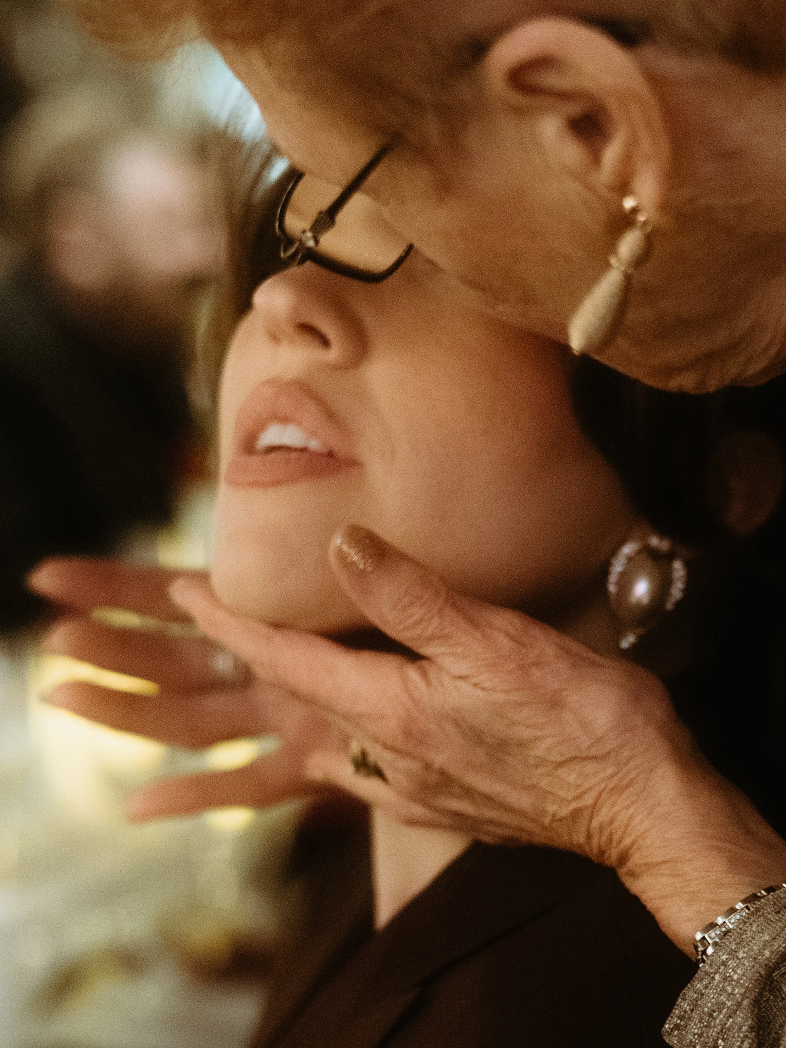 Close-up of an elderly woman and a younger woman with glasses sharing a moment, with the younger woman's face gently touching the elderly woman's hand.