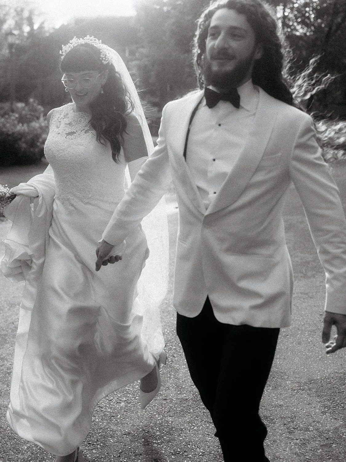 A newlywed couple holding hands and walking outdoors, the bride in a lace wedding dress with a veil and tiara, and the groom in a white tuxedo with a black bow tie.