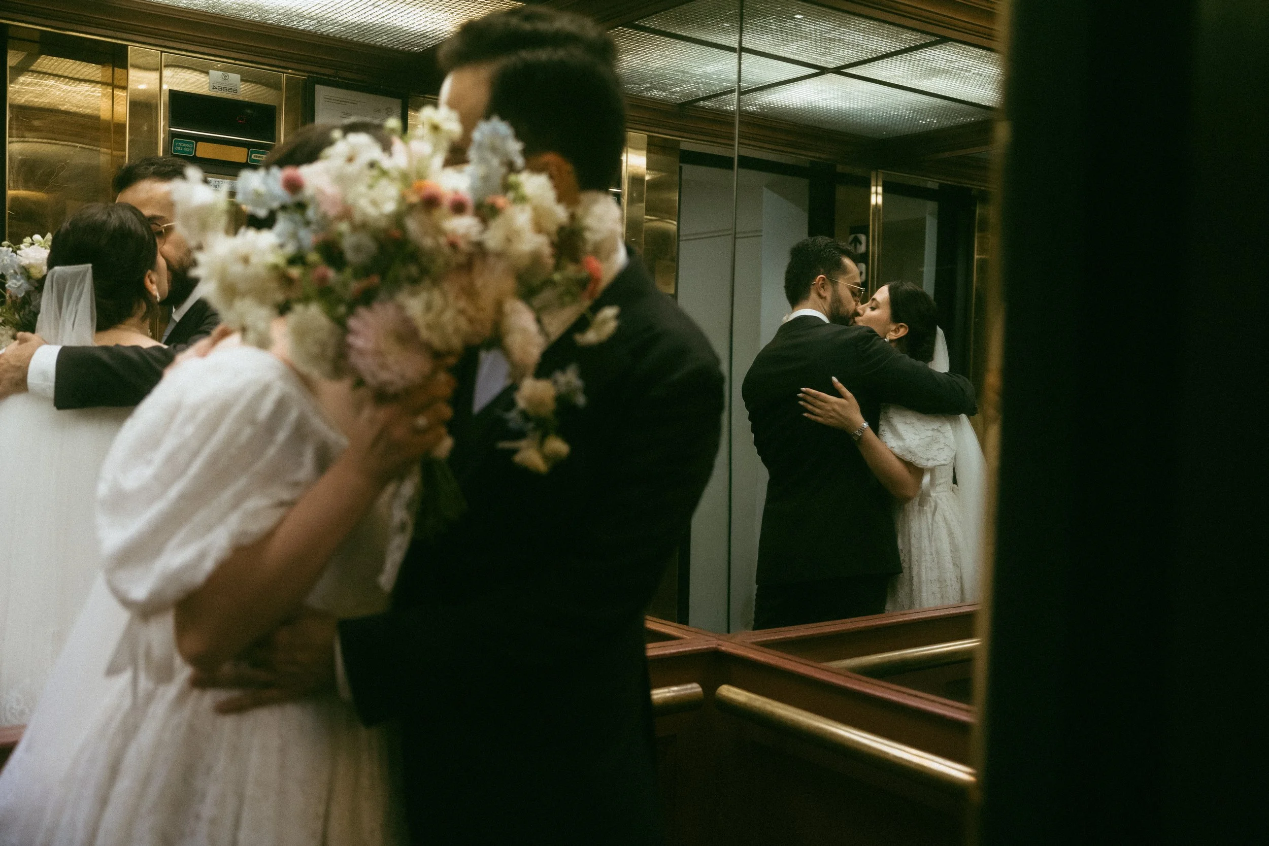 A wedding reception with multiple couples dancing closely, including a bride and groom sharing an embrace, seen through a mirror with the focus on the reflections, floral arrangements, and a warm, intimate ambiance.
