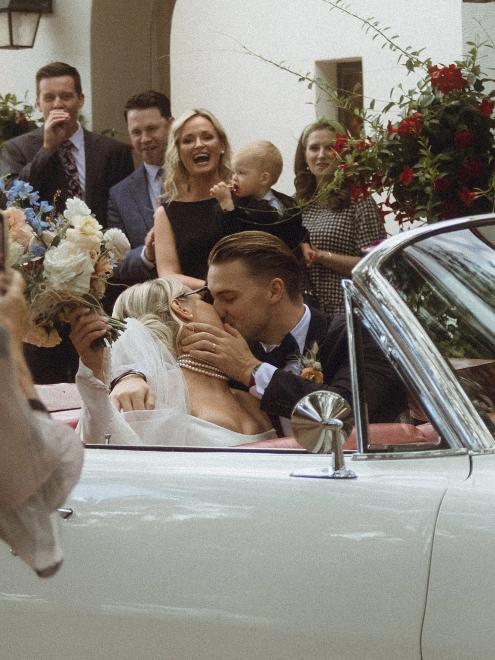 A newlywed couple sharing a kiss in a classic white convertible car, surrounded by wedding guests, some of whom are smiling and laughter, on a wedding day.