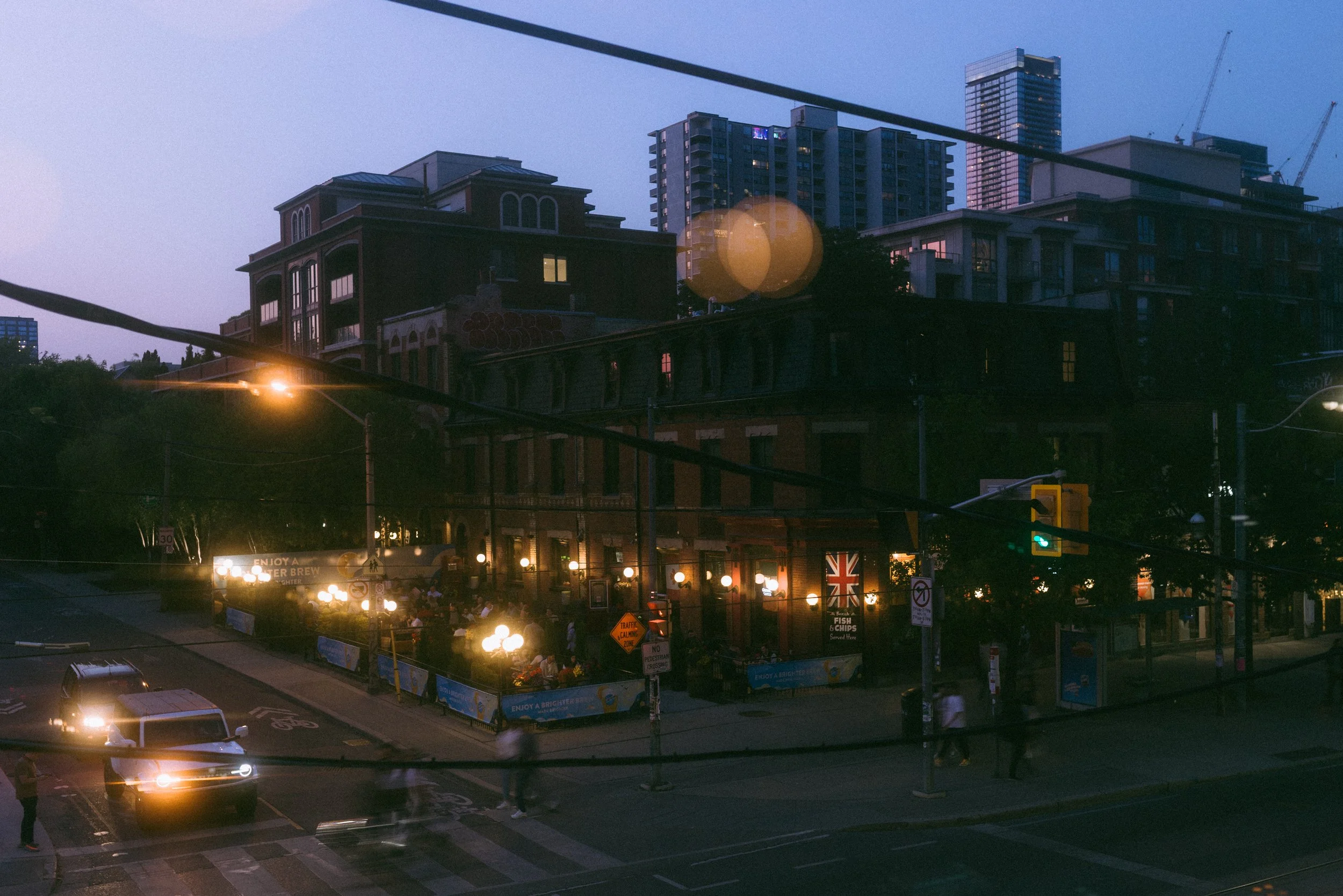 City street scene at dusk with buildings, streetlights, and cars. Pedestrians cross the intersection near a restaurant with outdoor seating illuminated by warm lights.