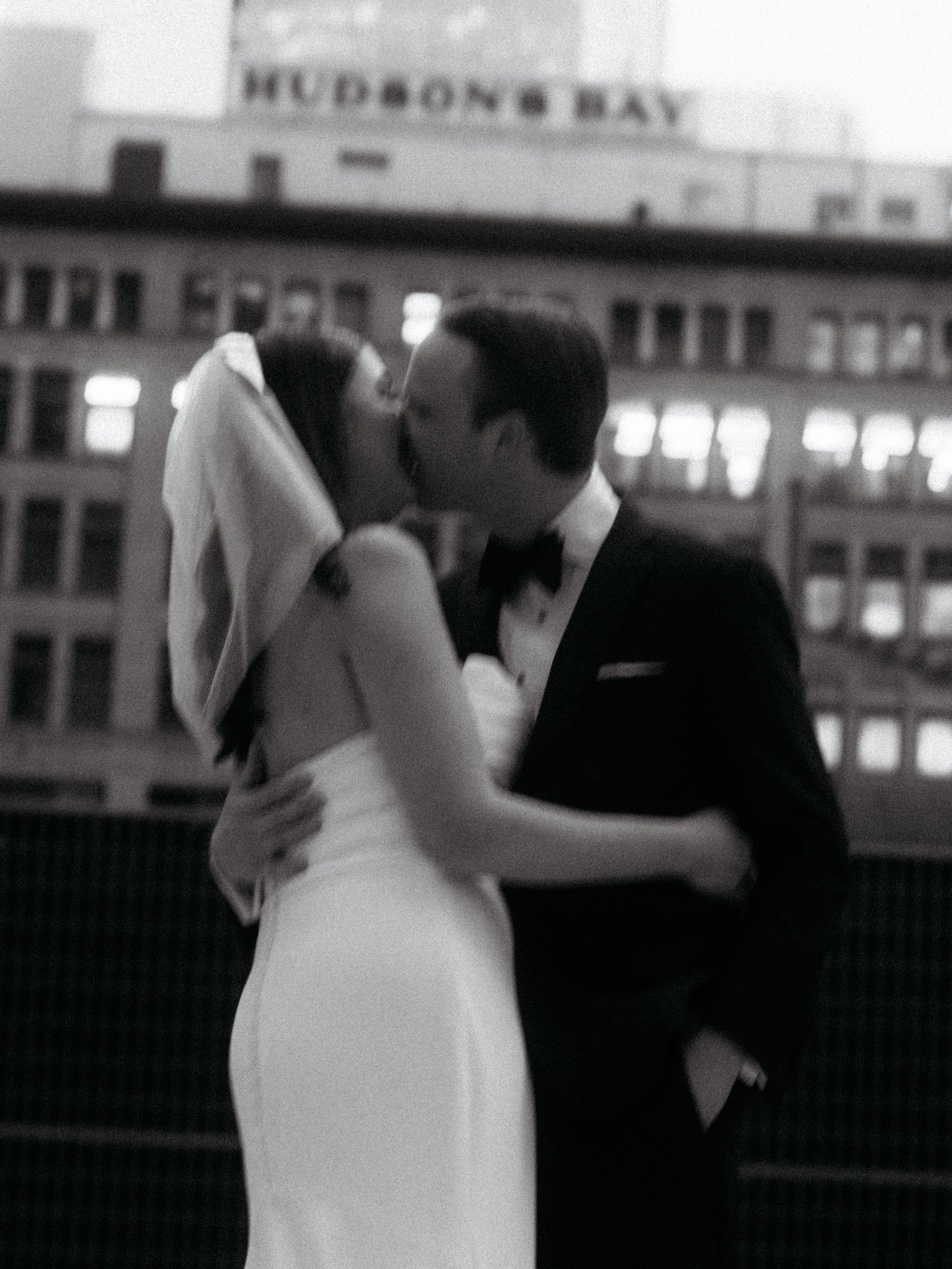 A couple in wedding attire sharing a kiss on a rooftop at night, with city buildings and a sign that reads 'HUDSON'S BAY' in the background.