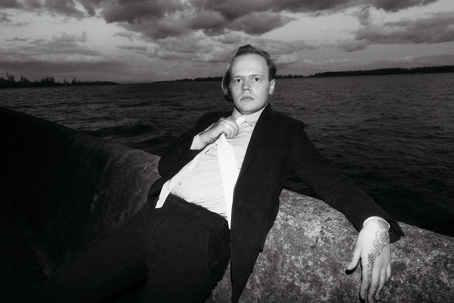A man in a suit reclining against a stone barrier by a body of water, with dark clouds in the sky.