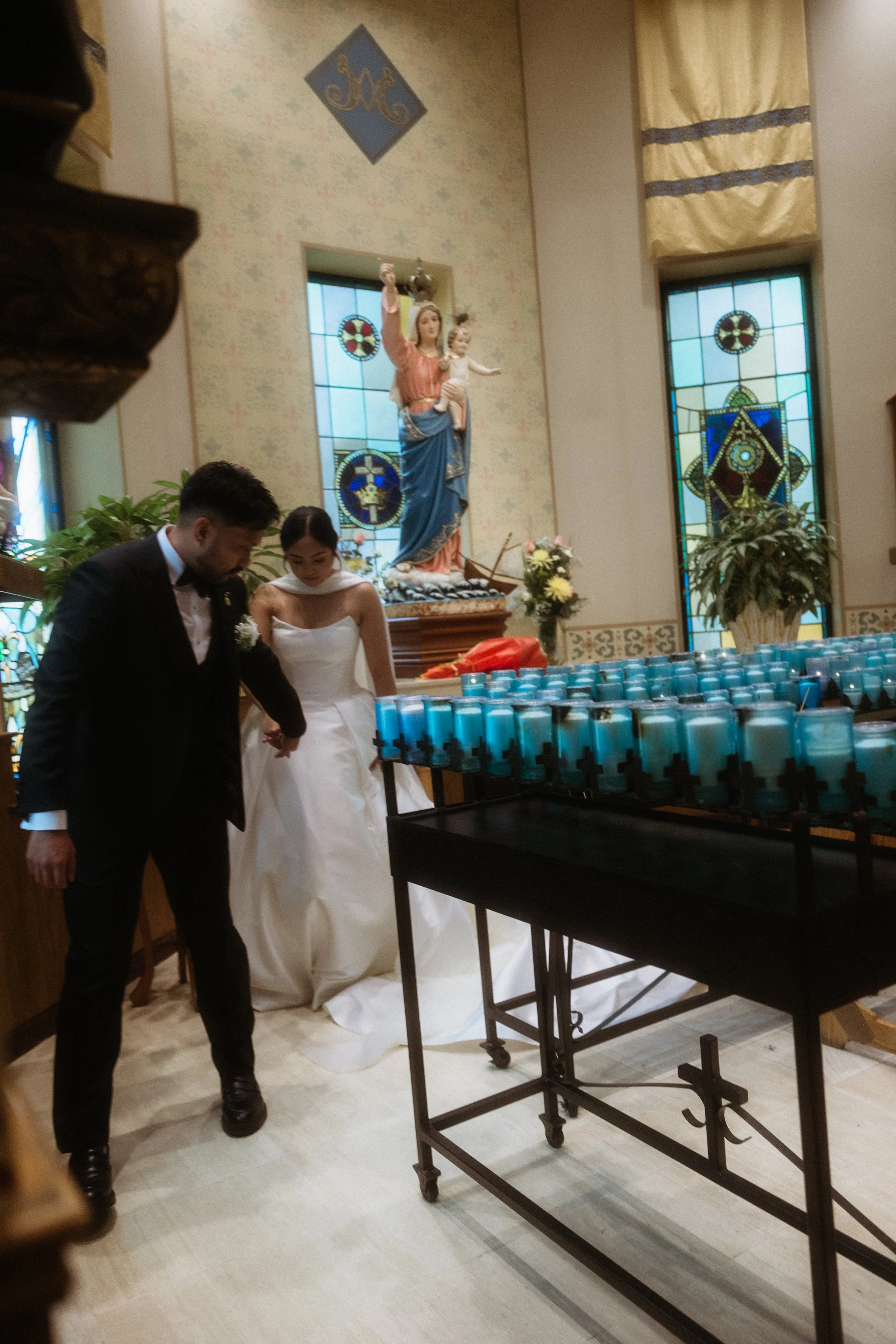 A newlywed couple in wedding attire inside a church, holding hands and walking towards the altar, with a large statue of Mary and Jesus and stained glass windows in the background.