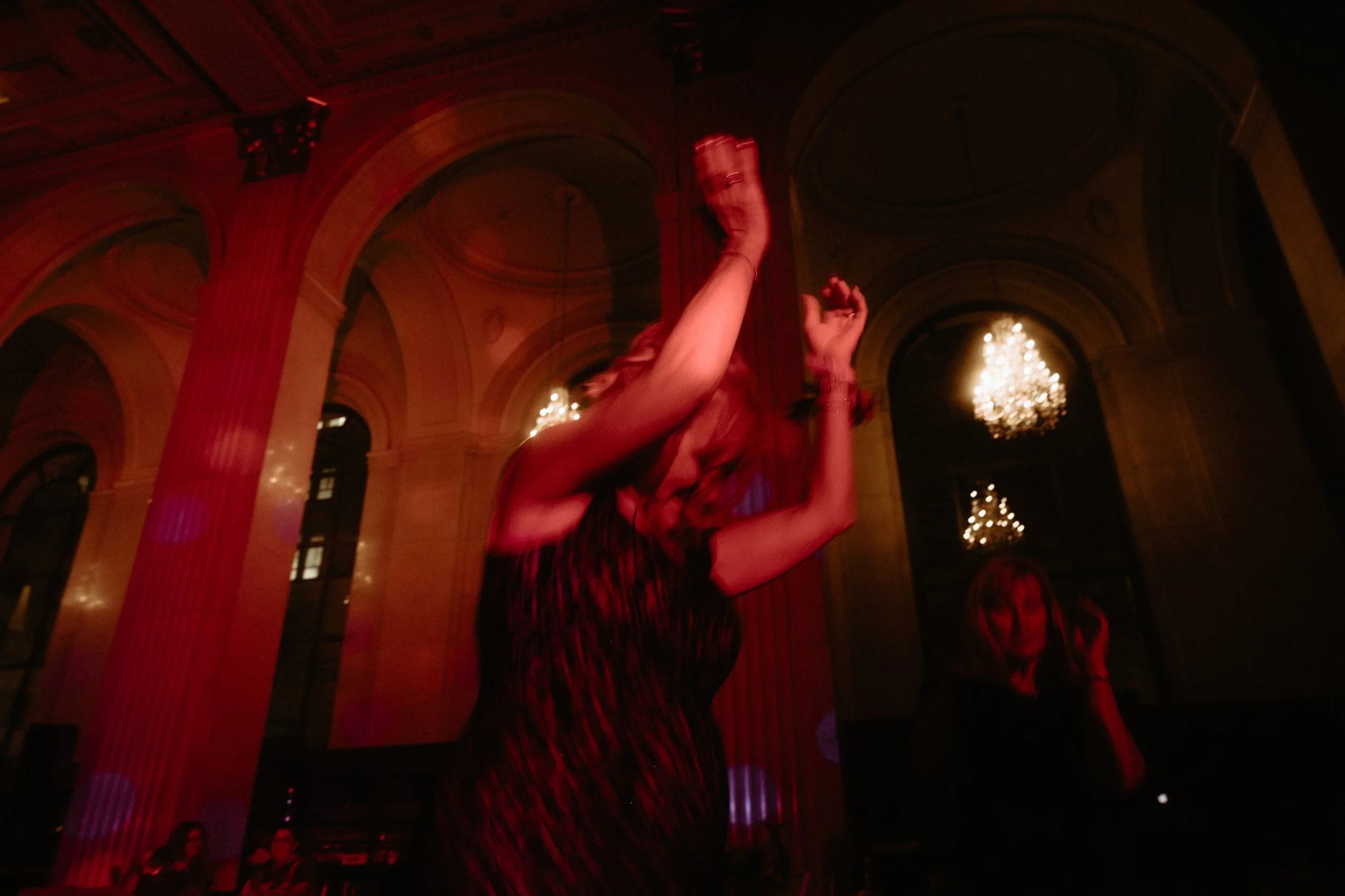 A woman dancing at a party or nightclub with chandeliers and Baroque-style architecture in the background, illuminated by red lighting.