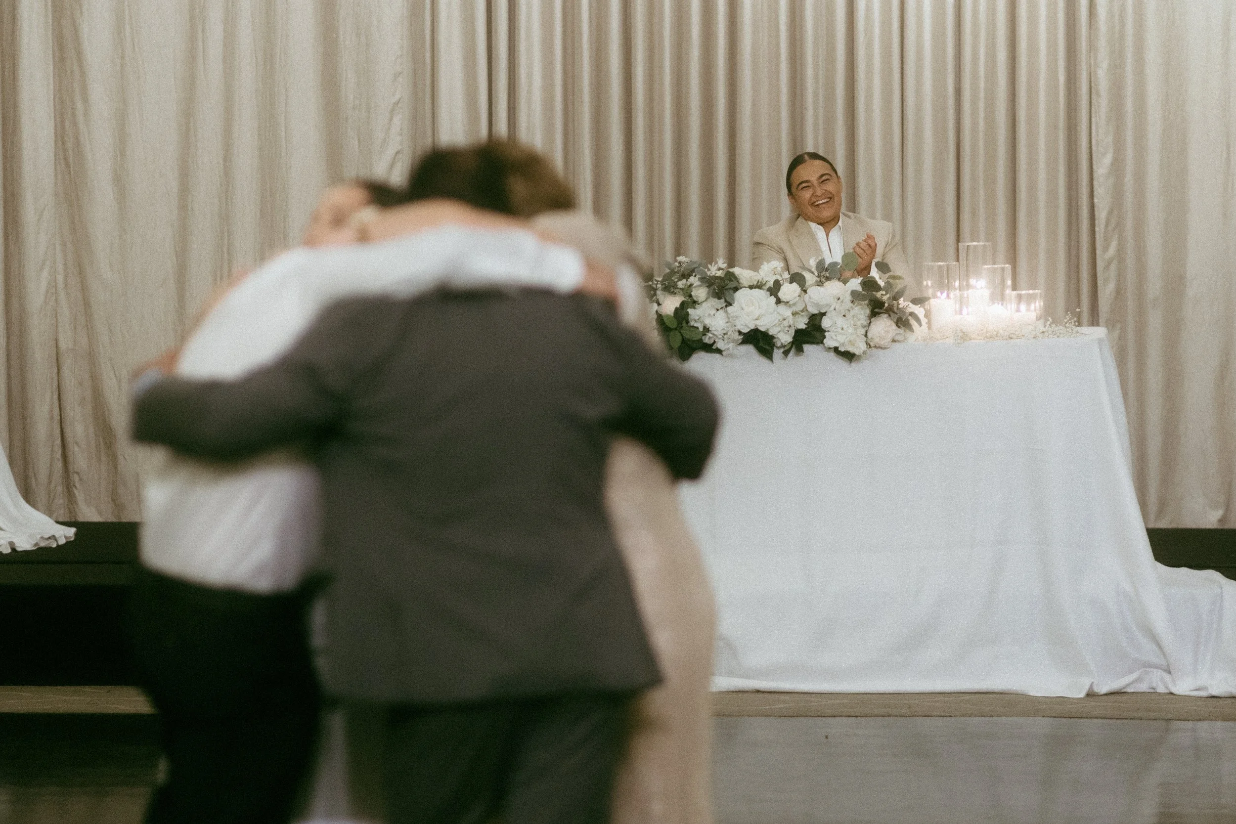 A bride and groom sharing a hug during their wedding ceremony with a female officiant sitting at a table decorated with flowers and candles in the background.