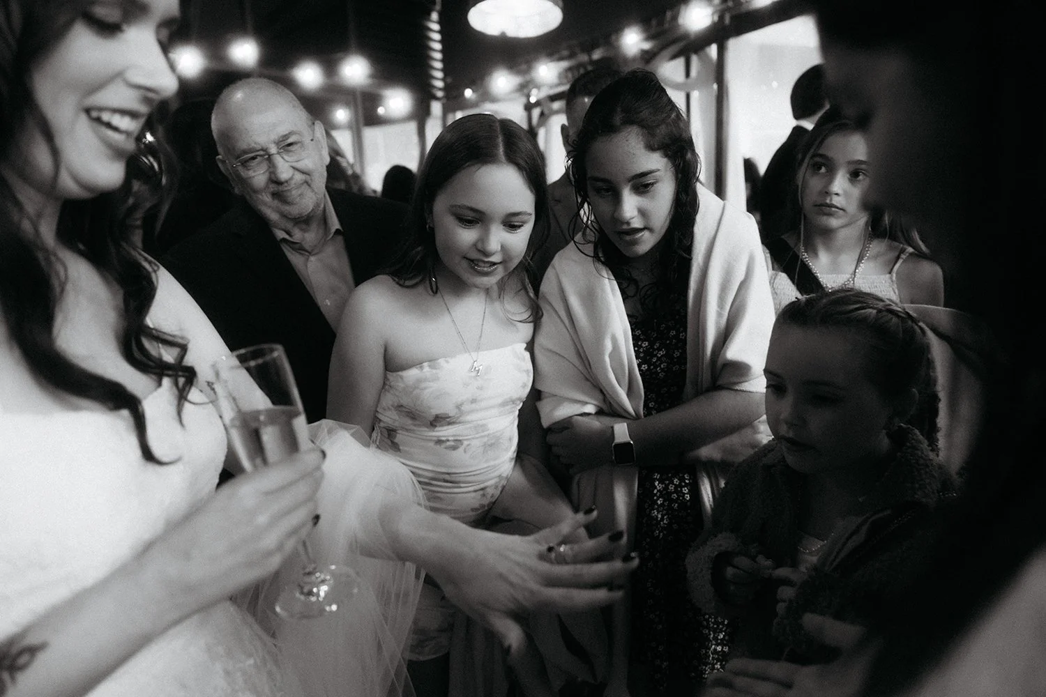 Group of women and children gathered together, looking at something on a cell phone, in a social setting with lights overhead