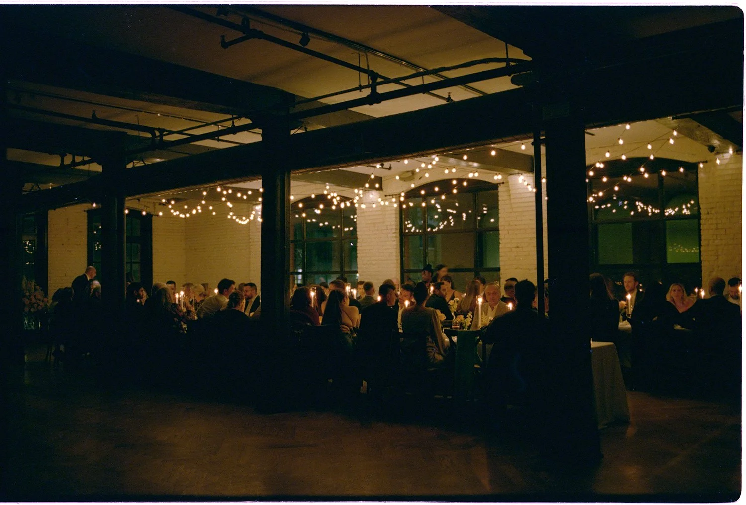 People sitting at tables in a dimly lit event space decorated with string lights, celebrating a formal gathering such as a wedding or party.