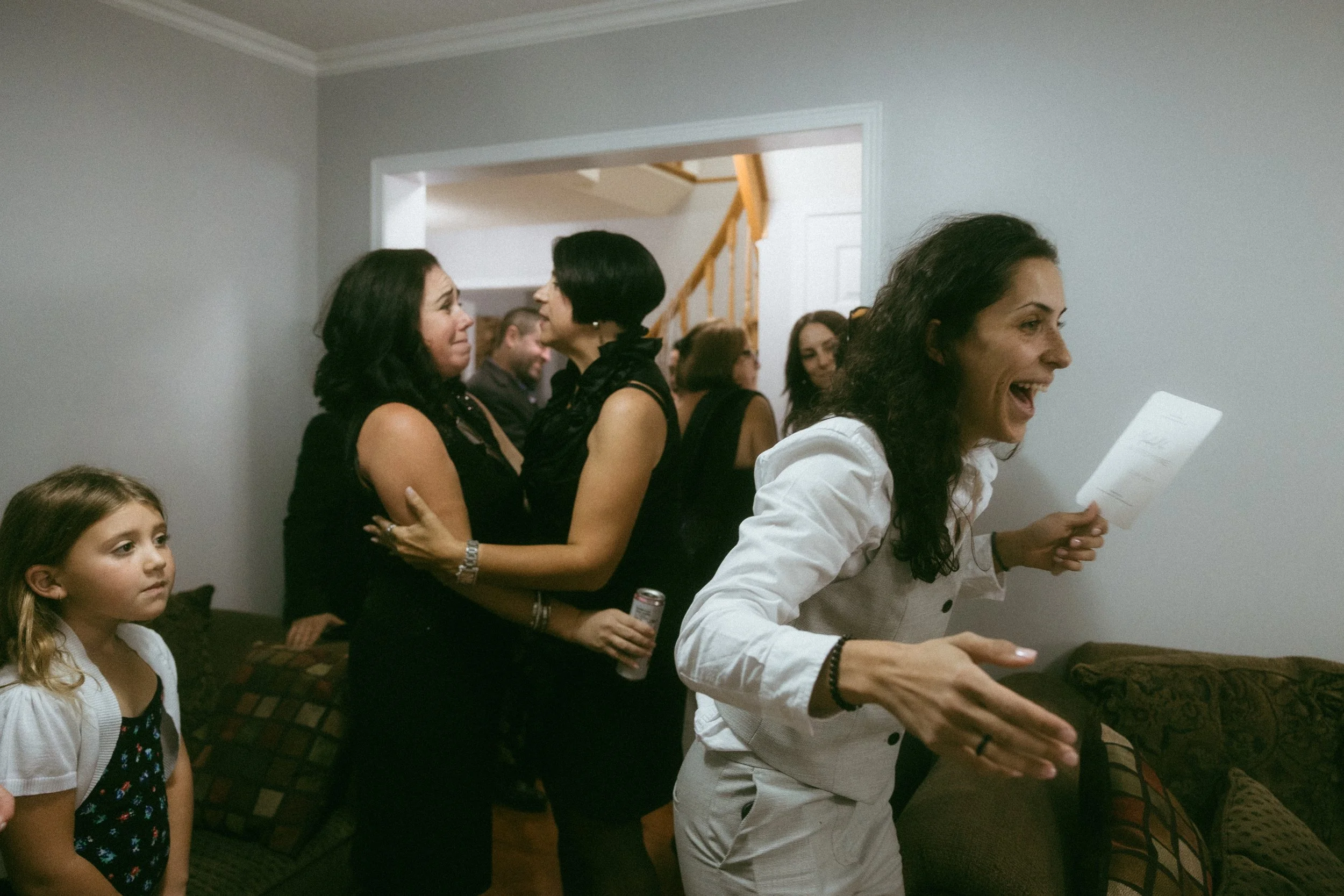A woman with dark curly hair wearing a white jacket excitedly greeting or talking to someone in a living room with several other people gathered, including two women embracing, a young girl standing nearby, and a man in the background.