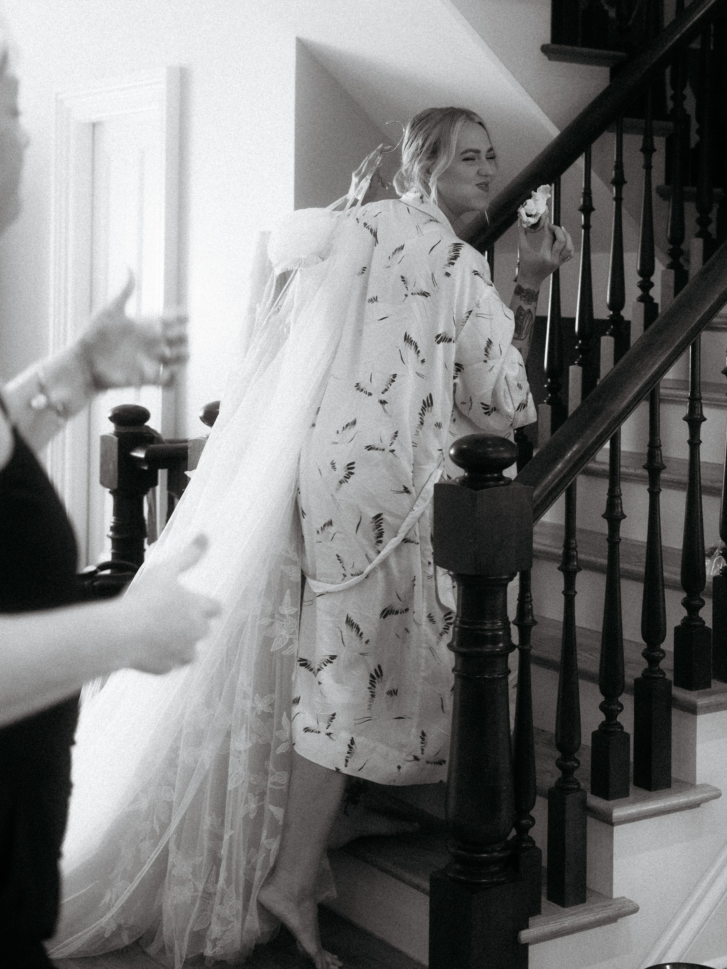 A woman in a patterned kimono holding a flower stands on a staircase, smiling at the camera. A person in the foreground is taking a photo or video.