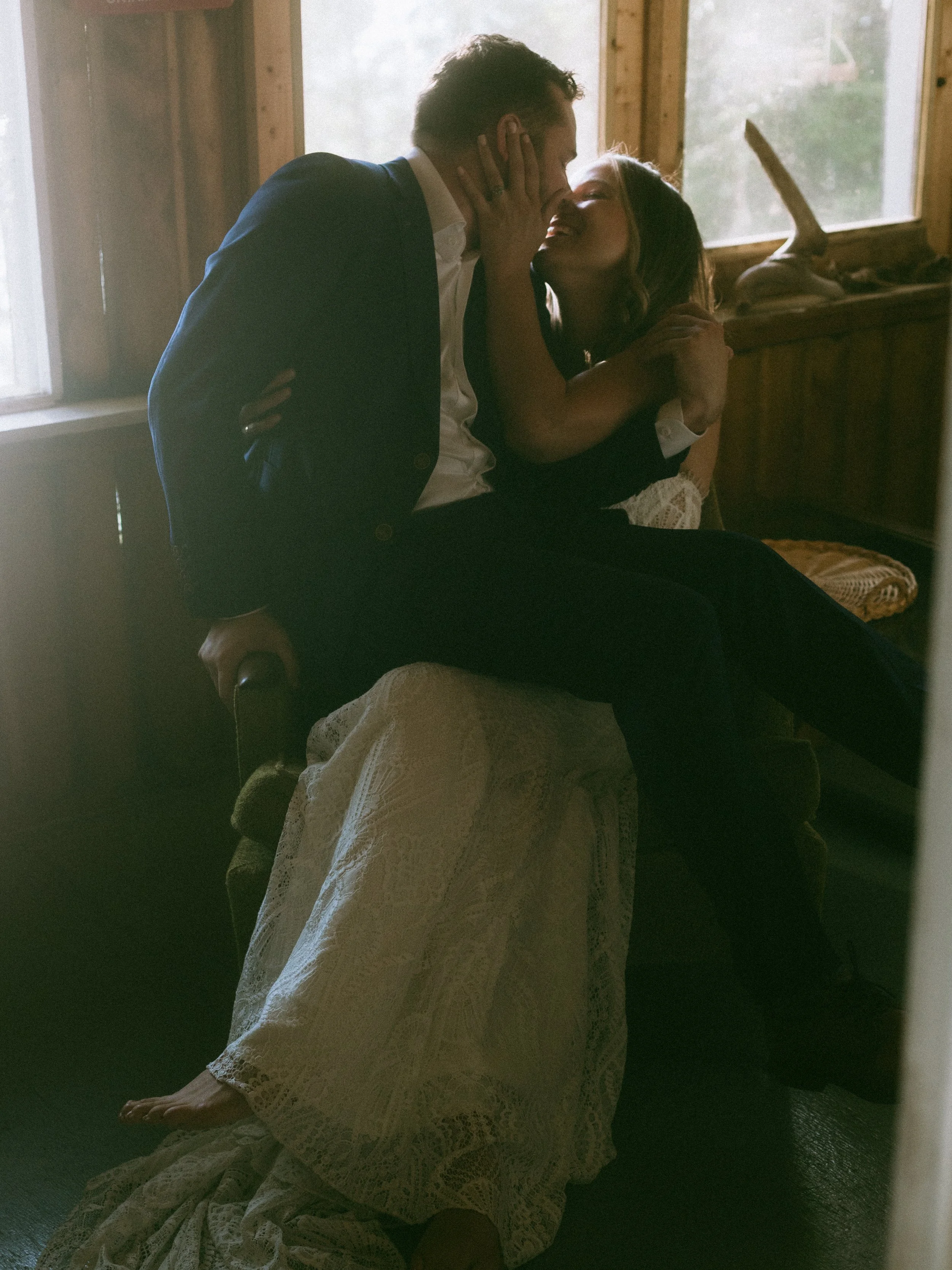 A couple sitting close together on a vintage armchair, sharing a kiss in a cozy, rustic wooden room with natural light coming through windows.