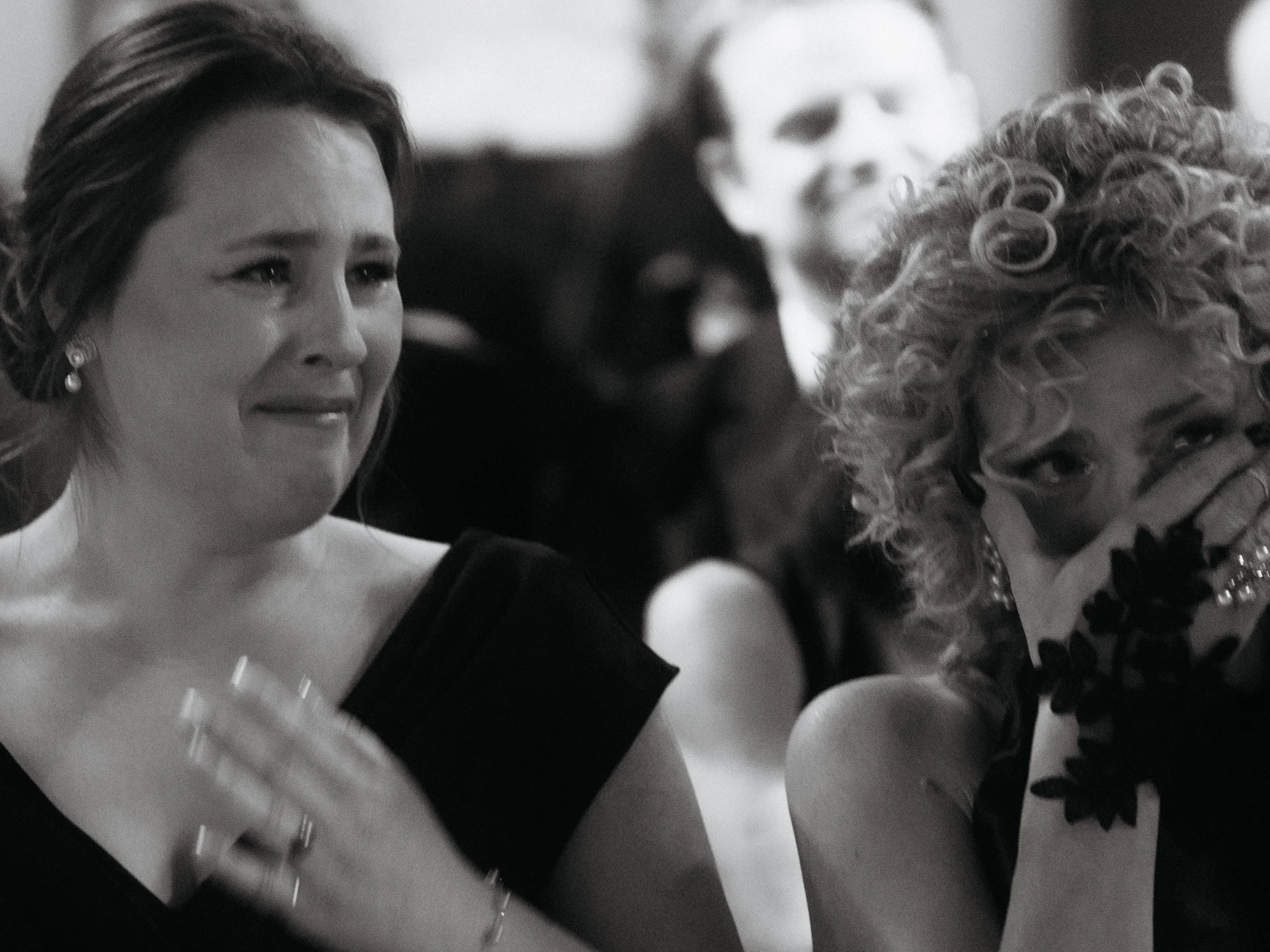 Two women at a formal event, sitting close together, one is crying and the other appears emotional, in black and white.