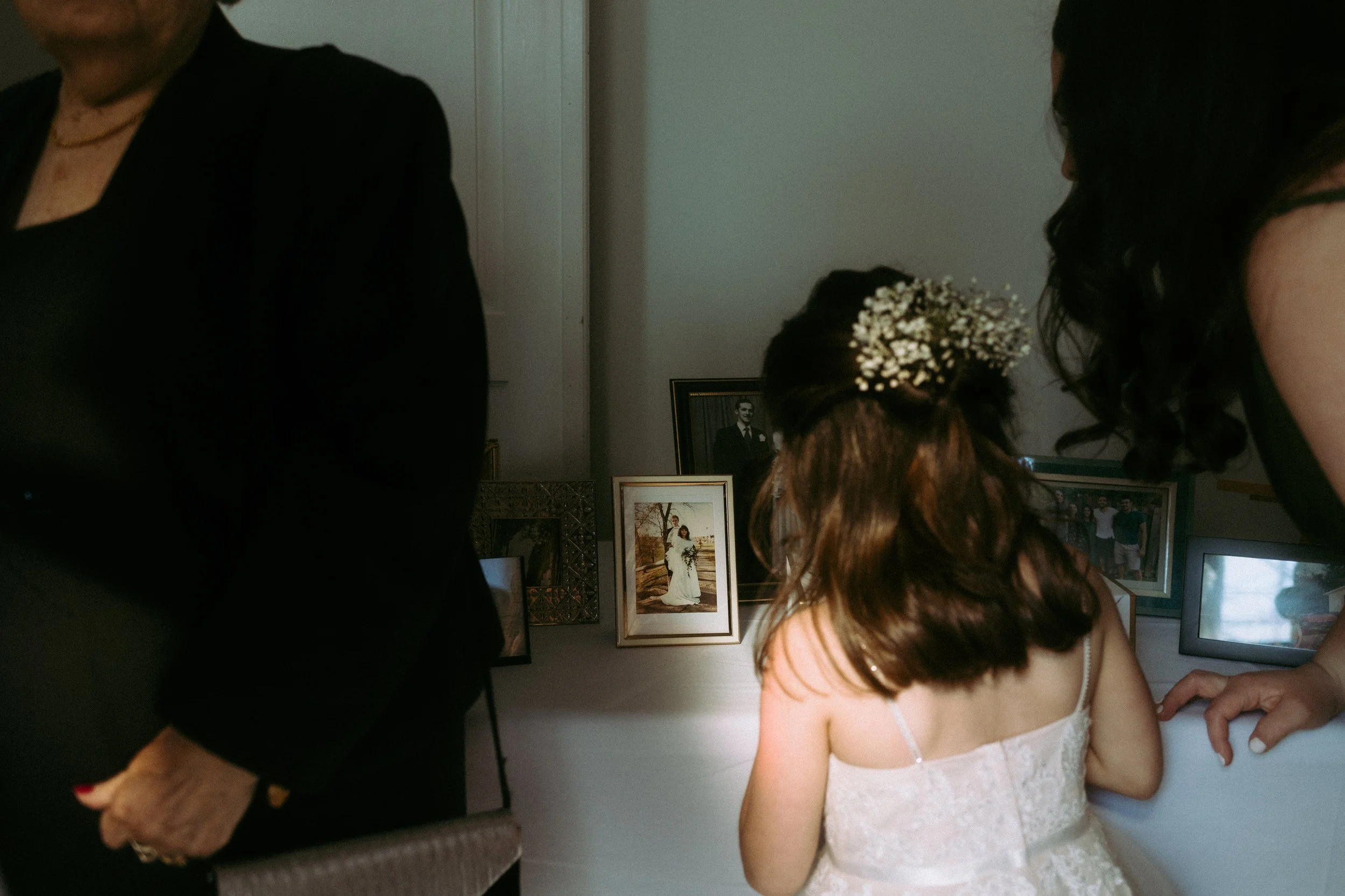 A young girl with brown hair adorned with small white flowers, wearing a white dress, looks at framed photographs displayed on a wall or table, with two partially visible women nearby.