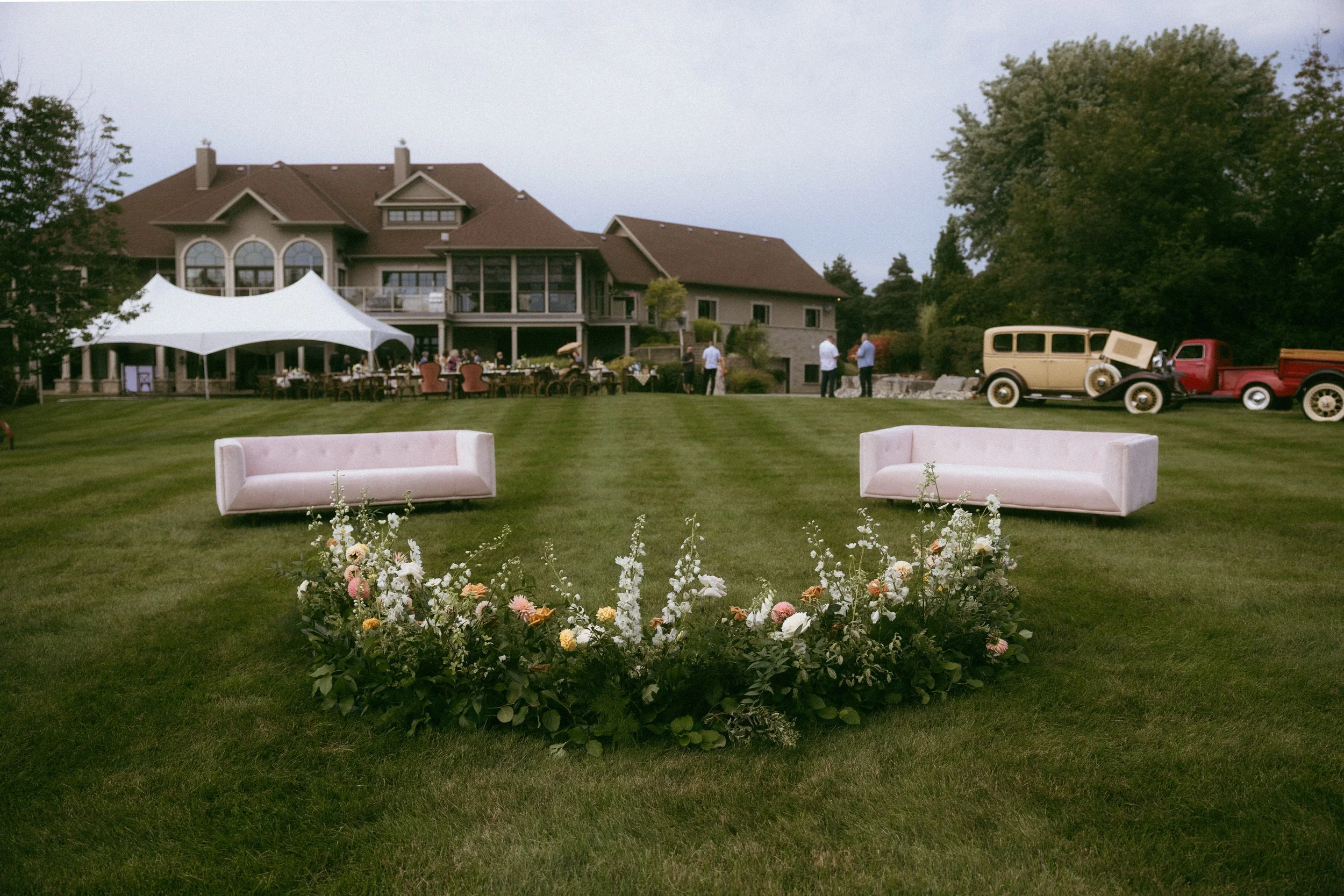 Outdoor wedding or event setup on a large lawn with vintage cars, pink sofas, a floral arrangement, and a large house with a tent and tables in the background.