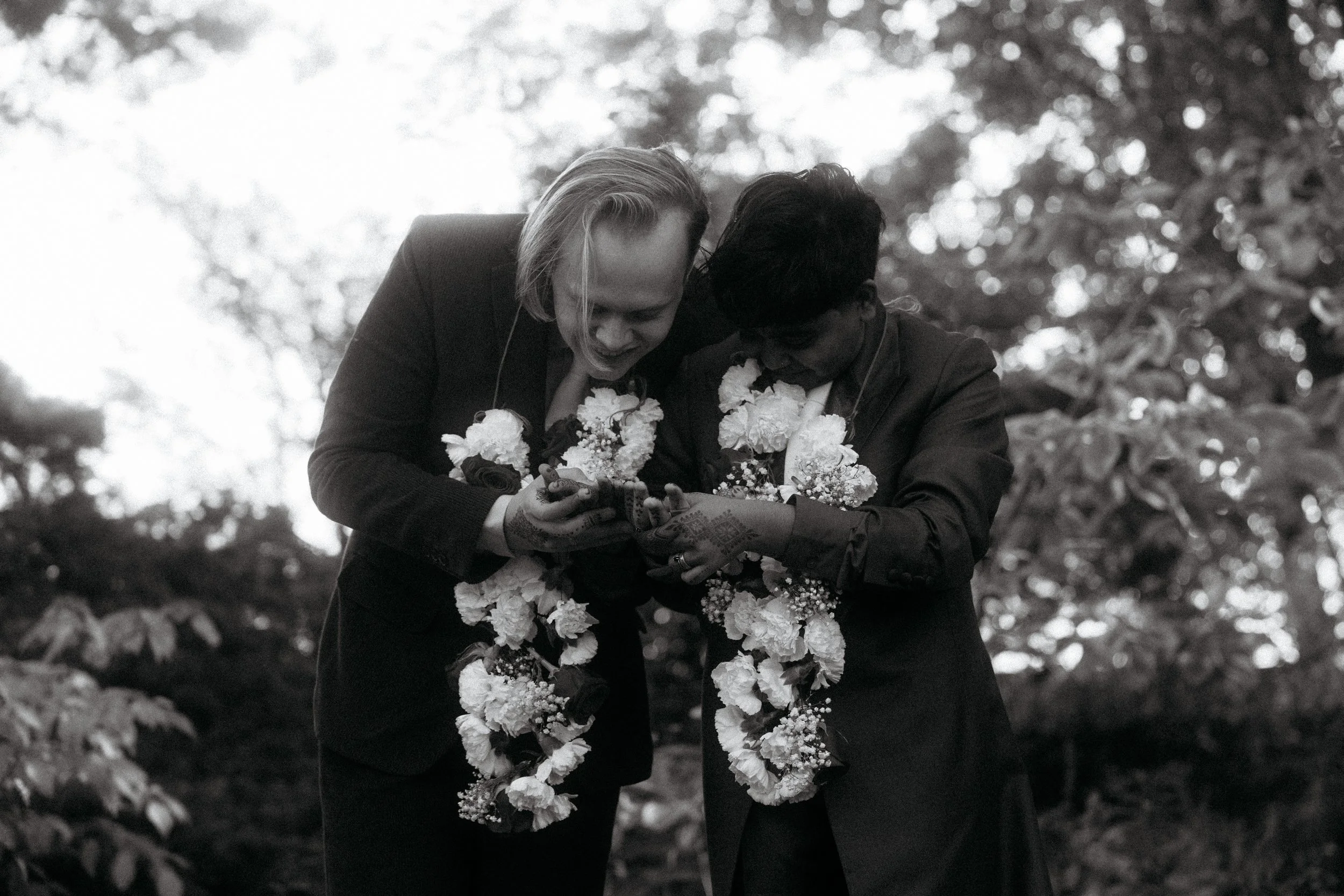 Two people in formal attire, one with lighter hair and one with darker hair, standing outdoors surrounded by trees, looking at a phone together. They are adorned with flower garlands around their necks and have henna designs on their hands.