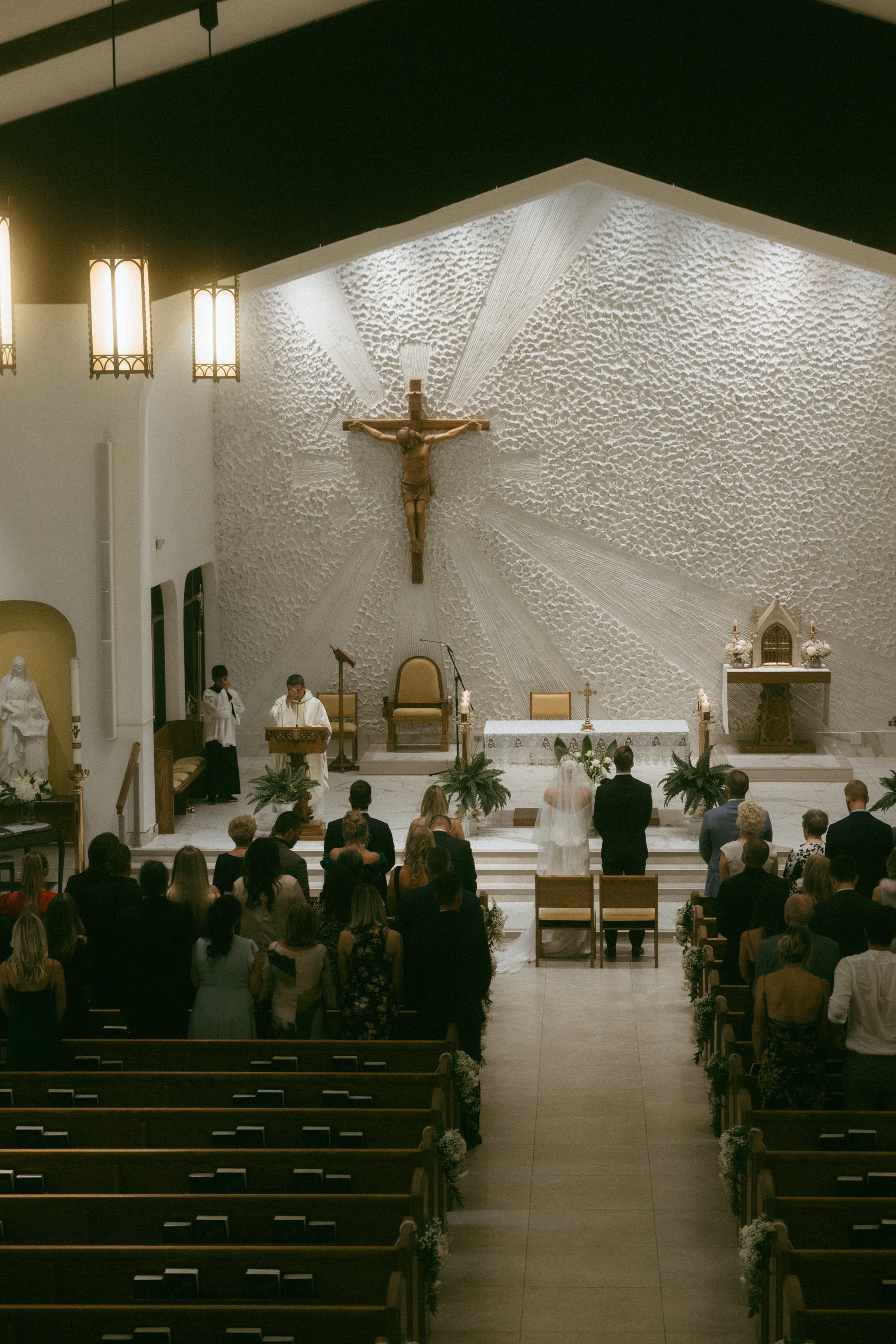 A wedding ceremony taking place inside a church with an altar and a large crucifix on a textured wall behind it, with guests standing and watching the couple.