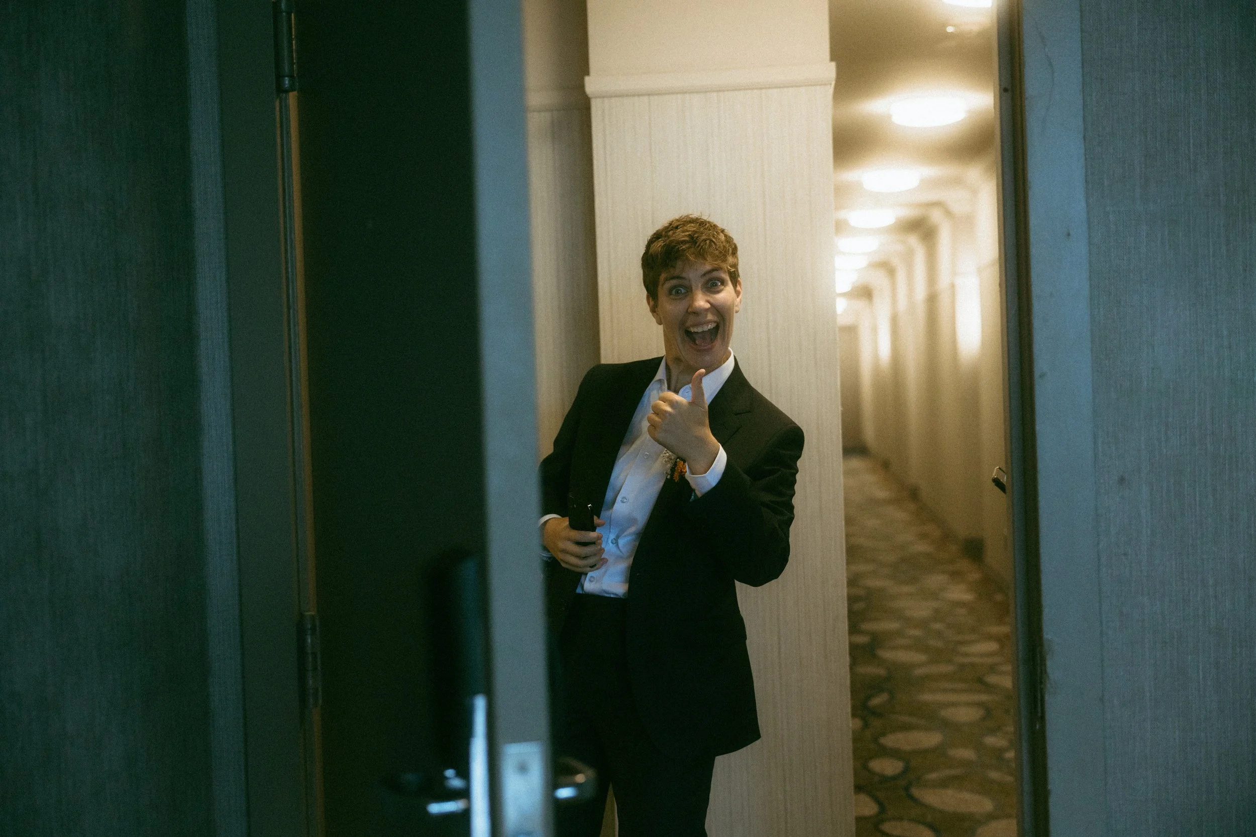 A person dressed in a tuxedo giving a thumbs up and smiling while standing in a hotel hallway.