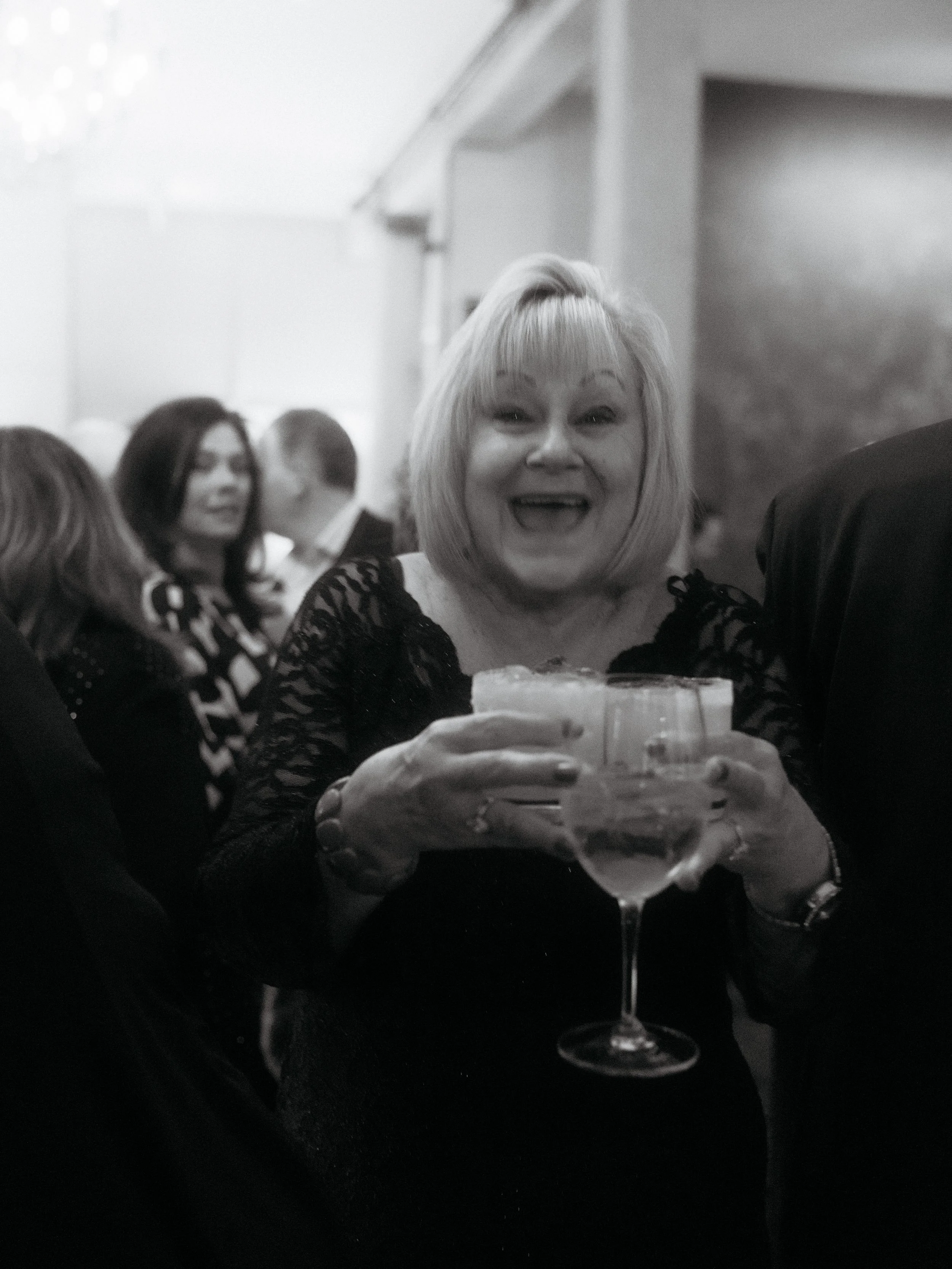 A woman with short blonde hair holding a glass of wine and smiling at a party or celebration.