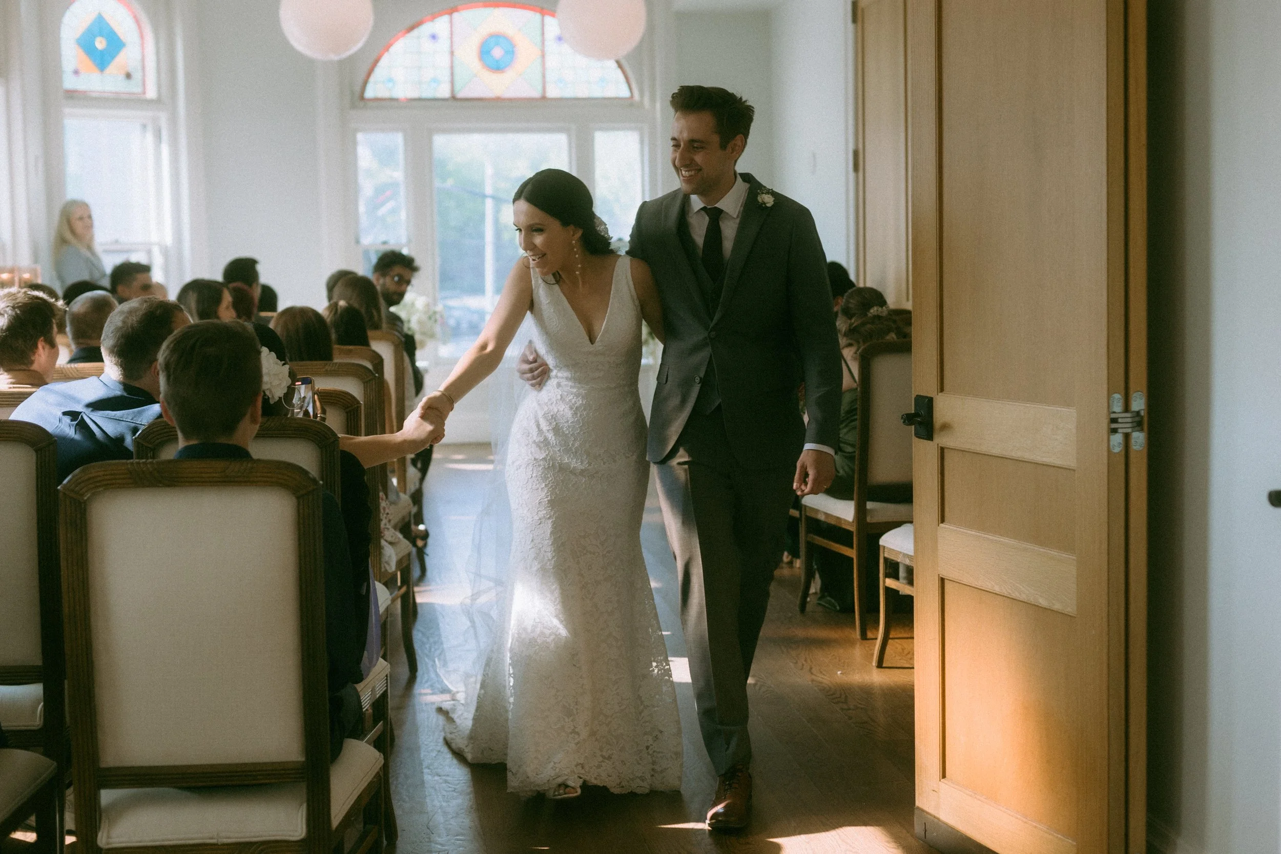 A bride and groom walking into a wedding ceremony, smiling and holding hands, with guests seated in the background inside a well-lit room with stained glass windows.