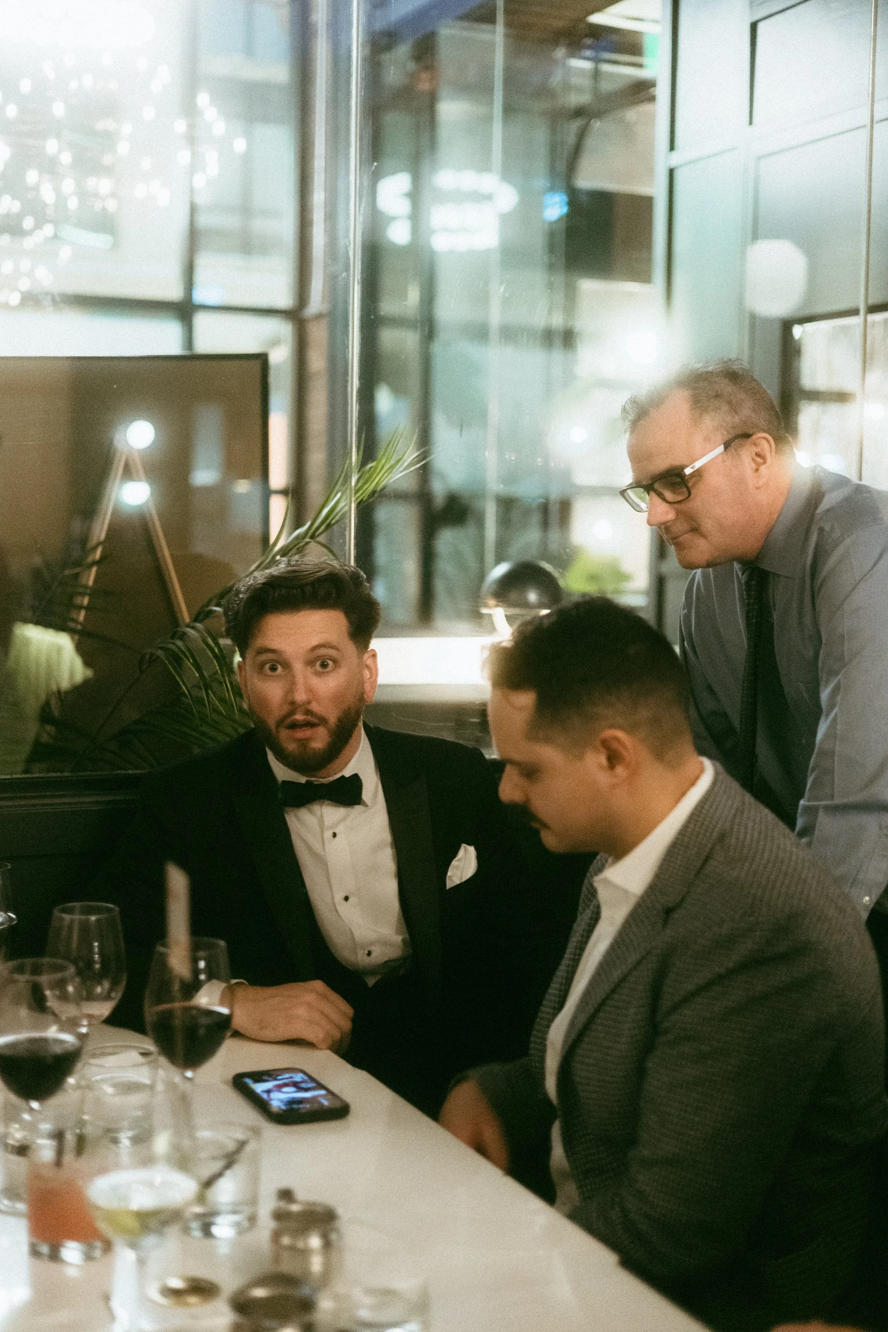 Three men in formal attire having a serious conversation at a restaurant table, with one man showing surprise and two others listening attentively.