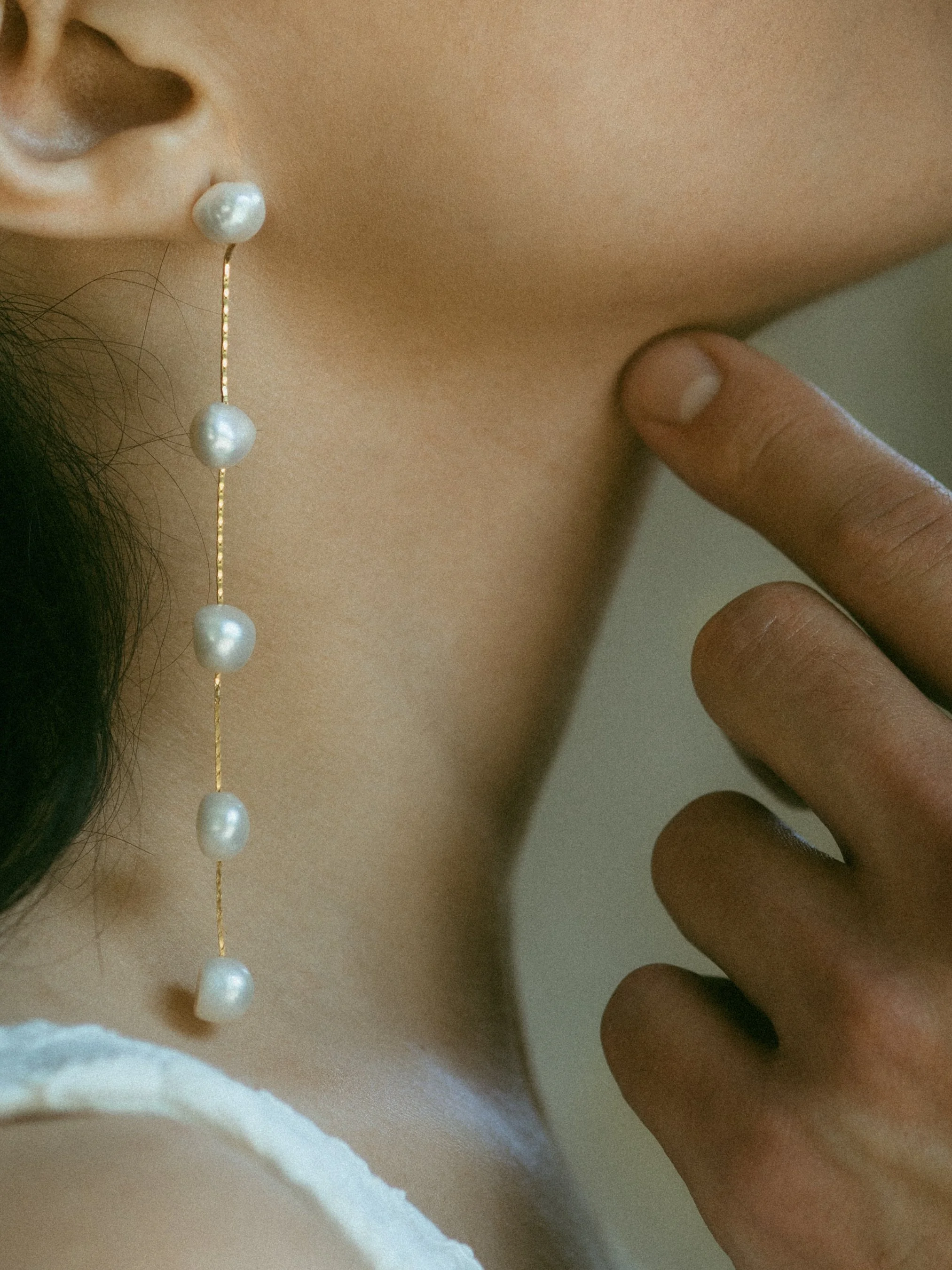Close-up of a woman's ear with a long pearl earring and her finger touching her neck.