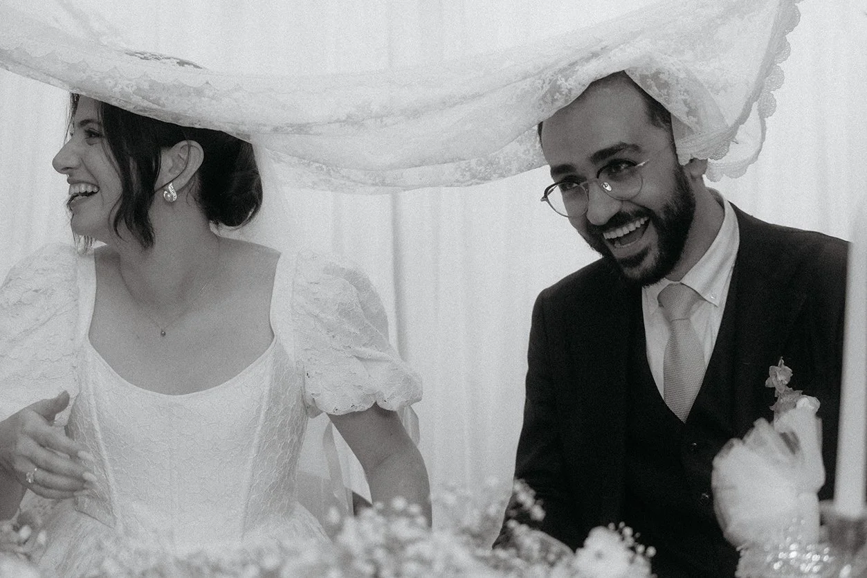 Black and white photo of a smiling bride and groom sitting at a wedding reception table, with the bride wearing a lace wedding dress and the groom in a suit and tie.