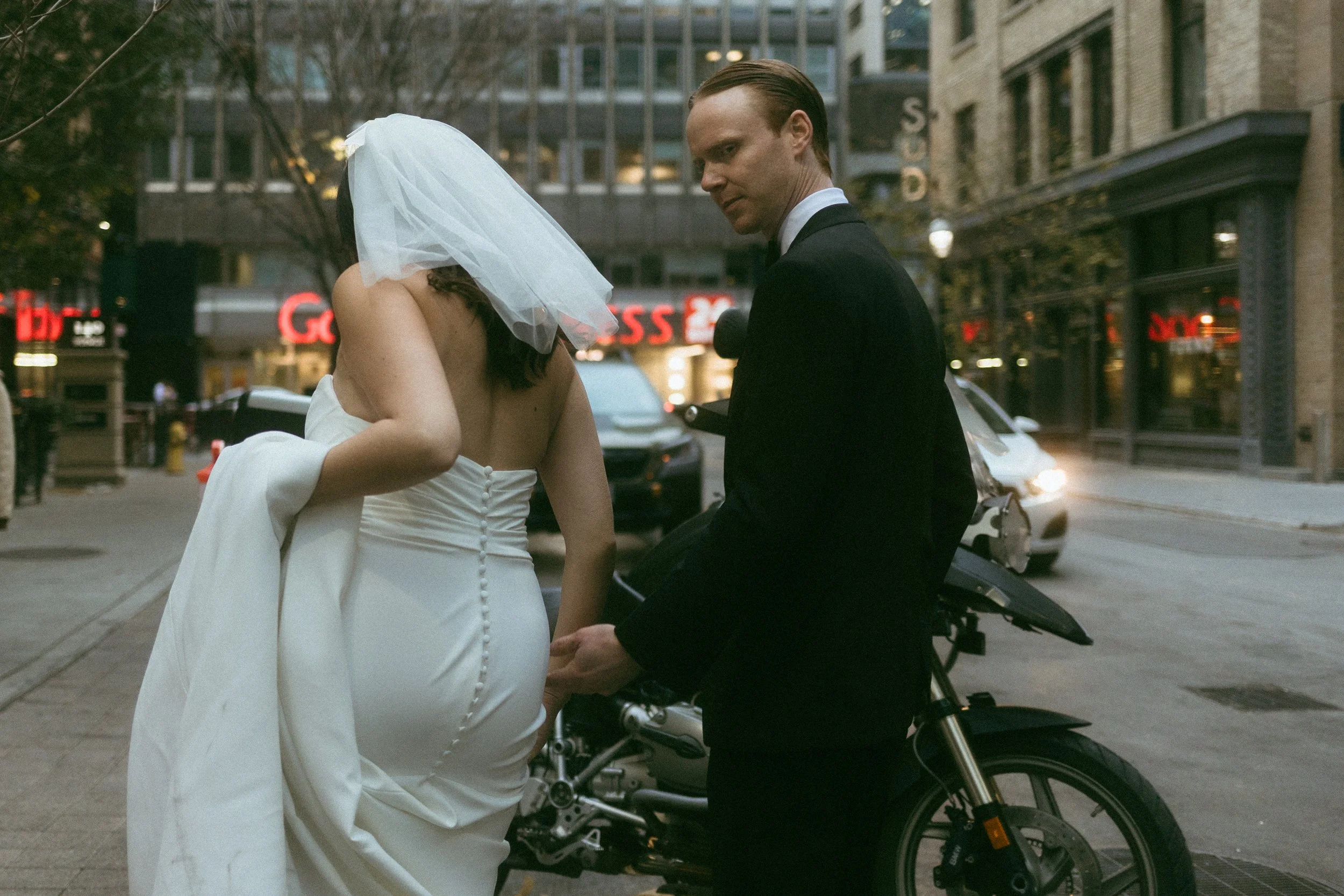 A bride in a white wedding dress and veil holding hands with a groom in a black suit, standing next to a motorcycle on a city street.