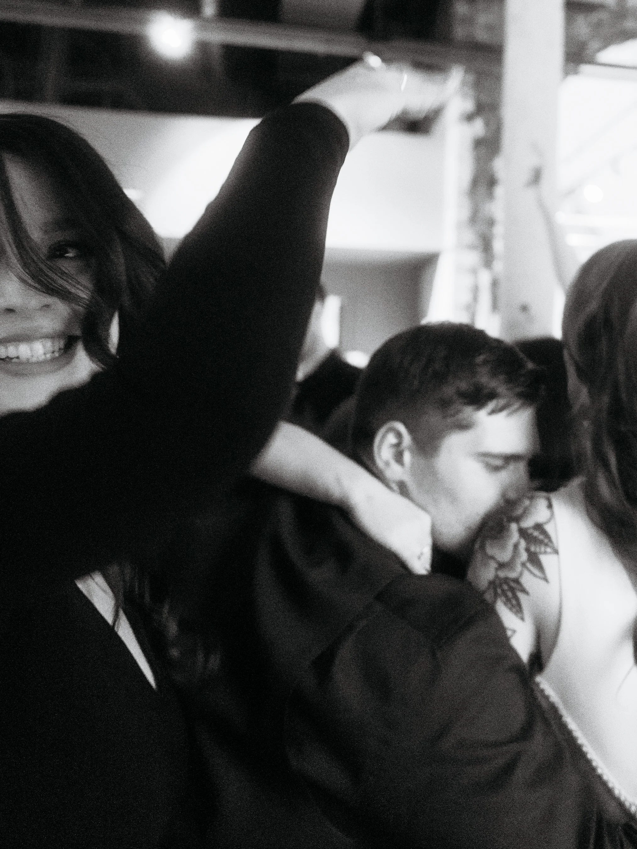 A black-and-white photo of people at a social gathering, with a smiling woman on the left and a woman with a tattooed shoulder near a young man. The woman on the left is smiling with her teeth visible.