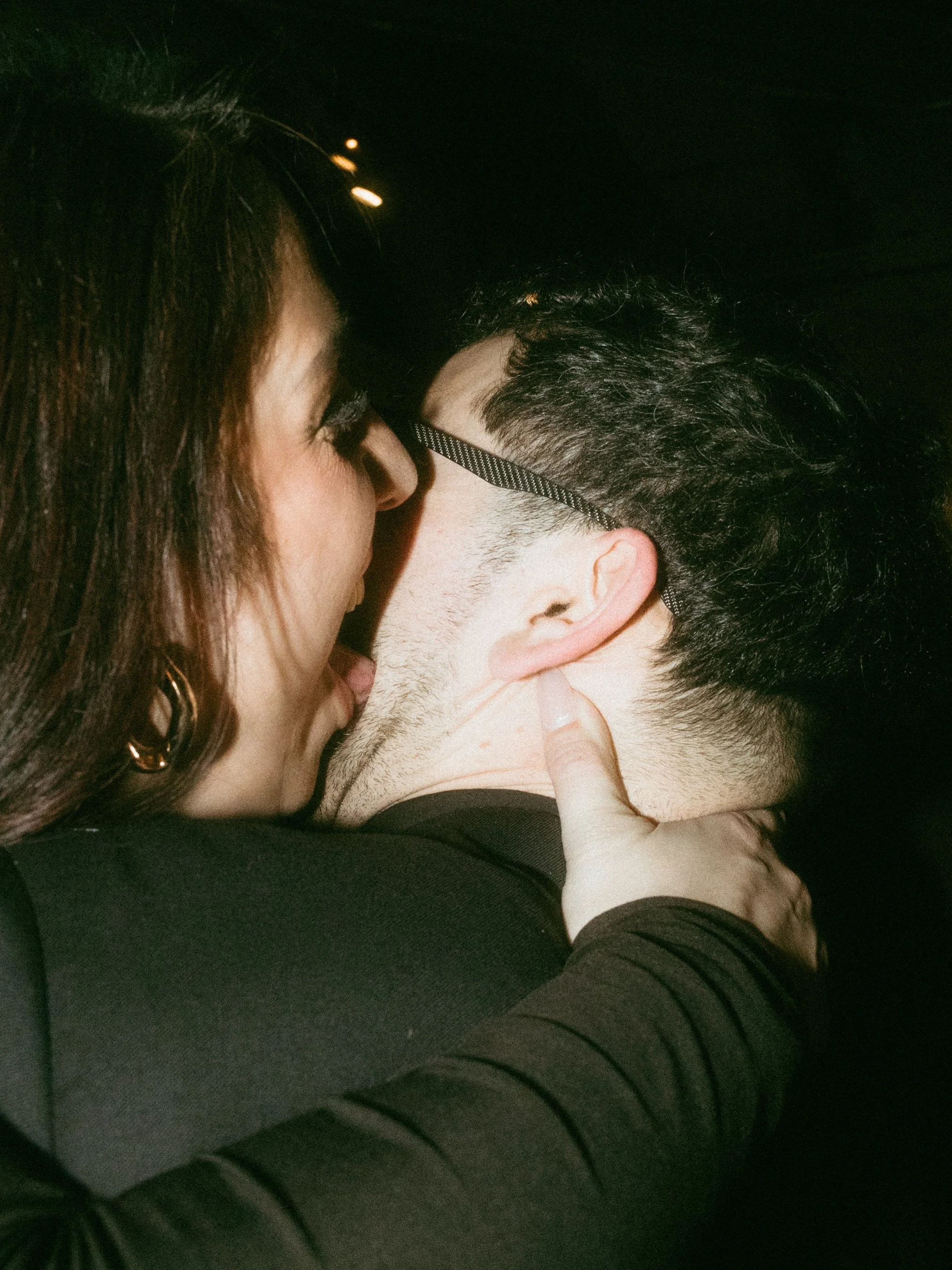 A woman and a man are close together, with the woman kissing the man's cheek as she holds his head. The woman has dark hair and is wearing earrings, while the man has glasses and dark hair. The scene appears to be in a dark environment, possibly at n