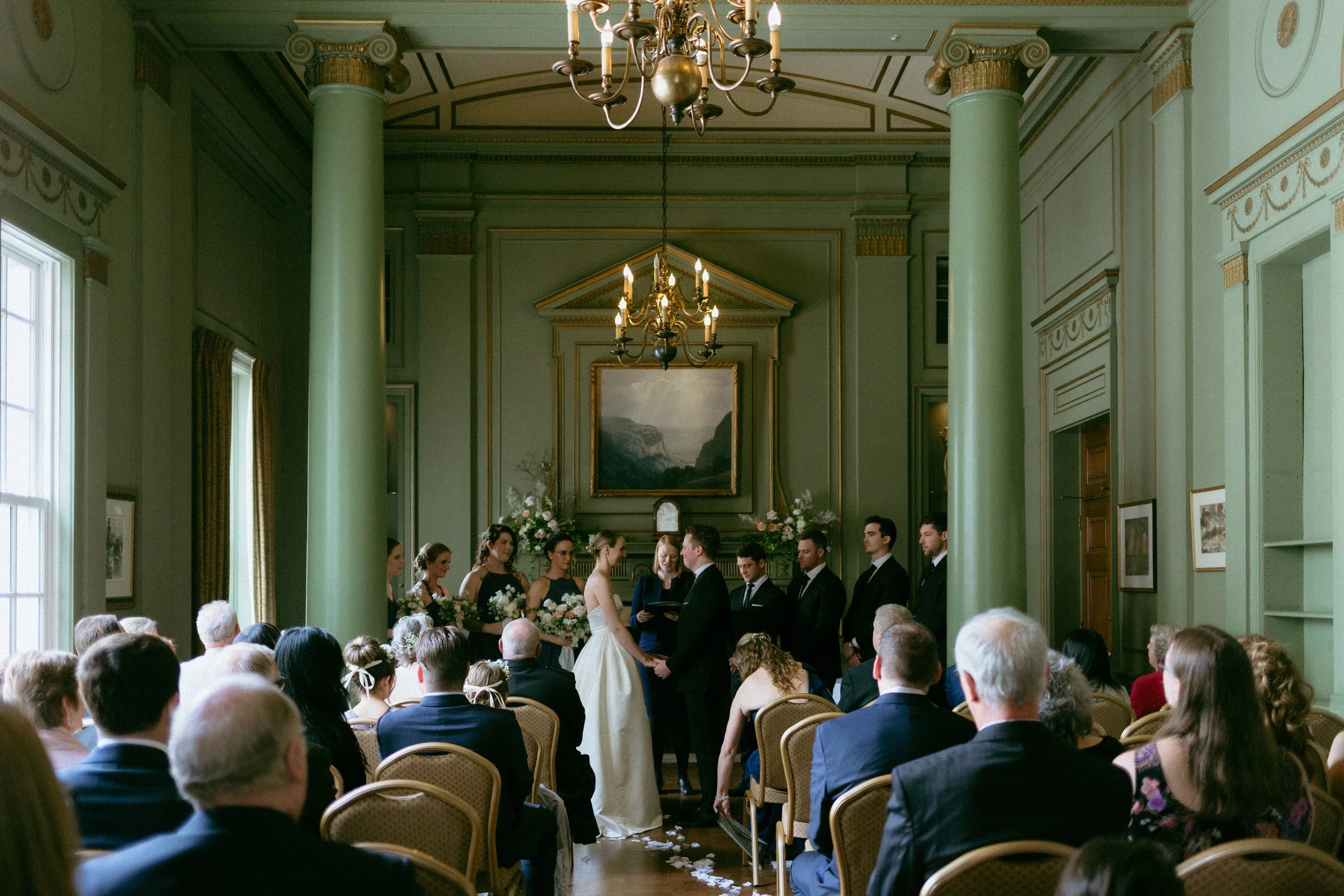 A wedding ceremony taking place in a grand, ornate room with high ceilings, green pillars, and large windows. The bride and groom stand facing each other at the altar, surrounded by bridesmaids and groomsmen, with guests seated in rows watching the c