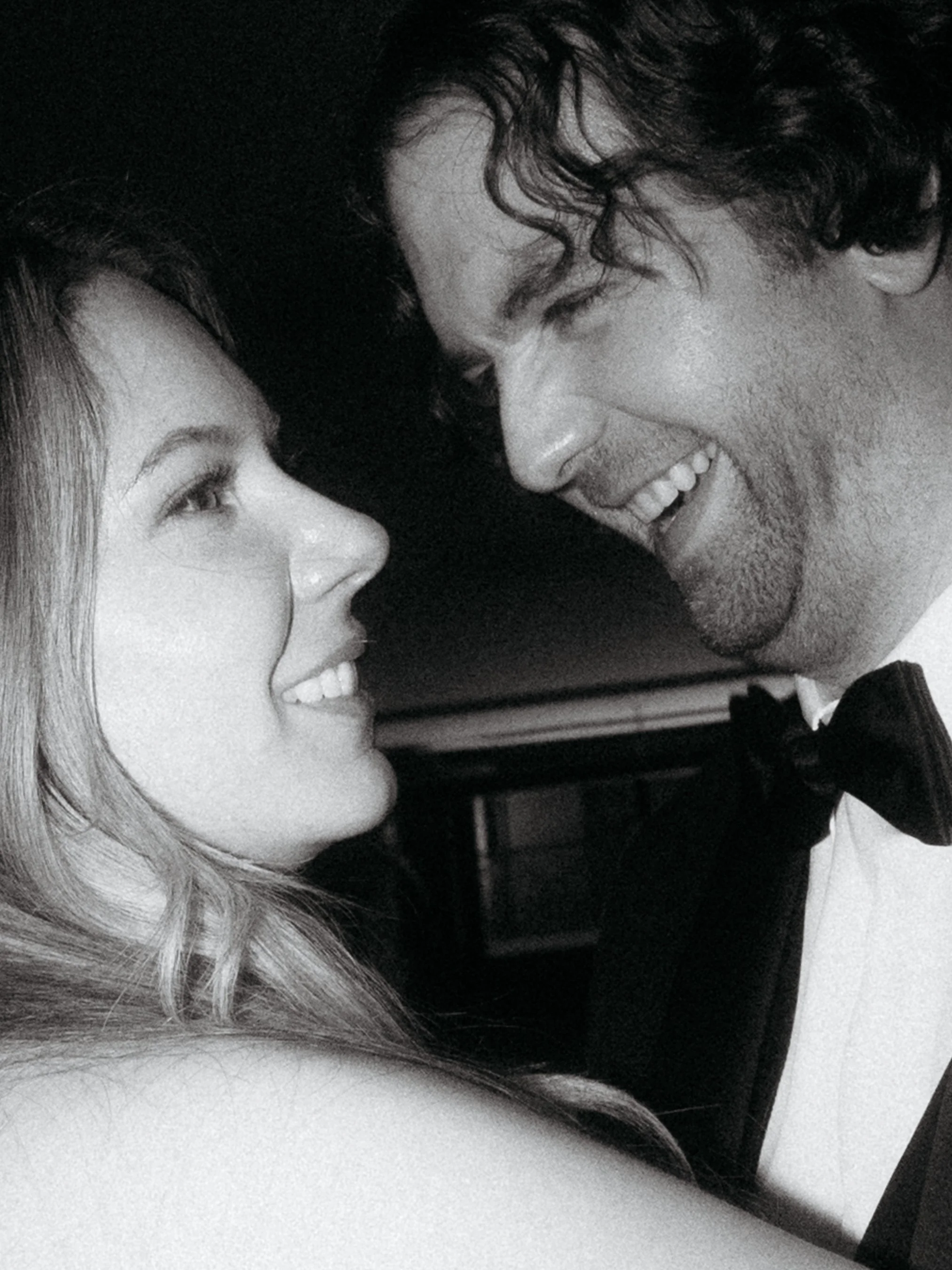 A black and white photo of a woman and a man smiling with noses nearly touching, dressed in formal attire, with the man wearing a tuxedo and bow tie.
