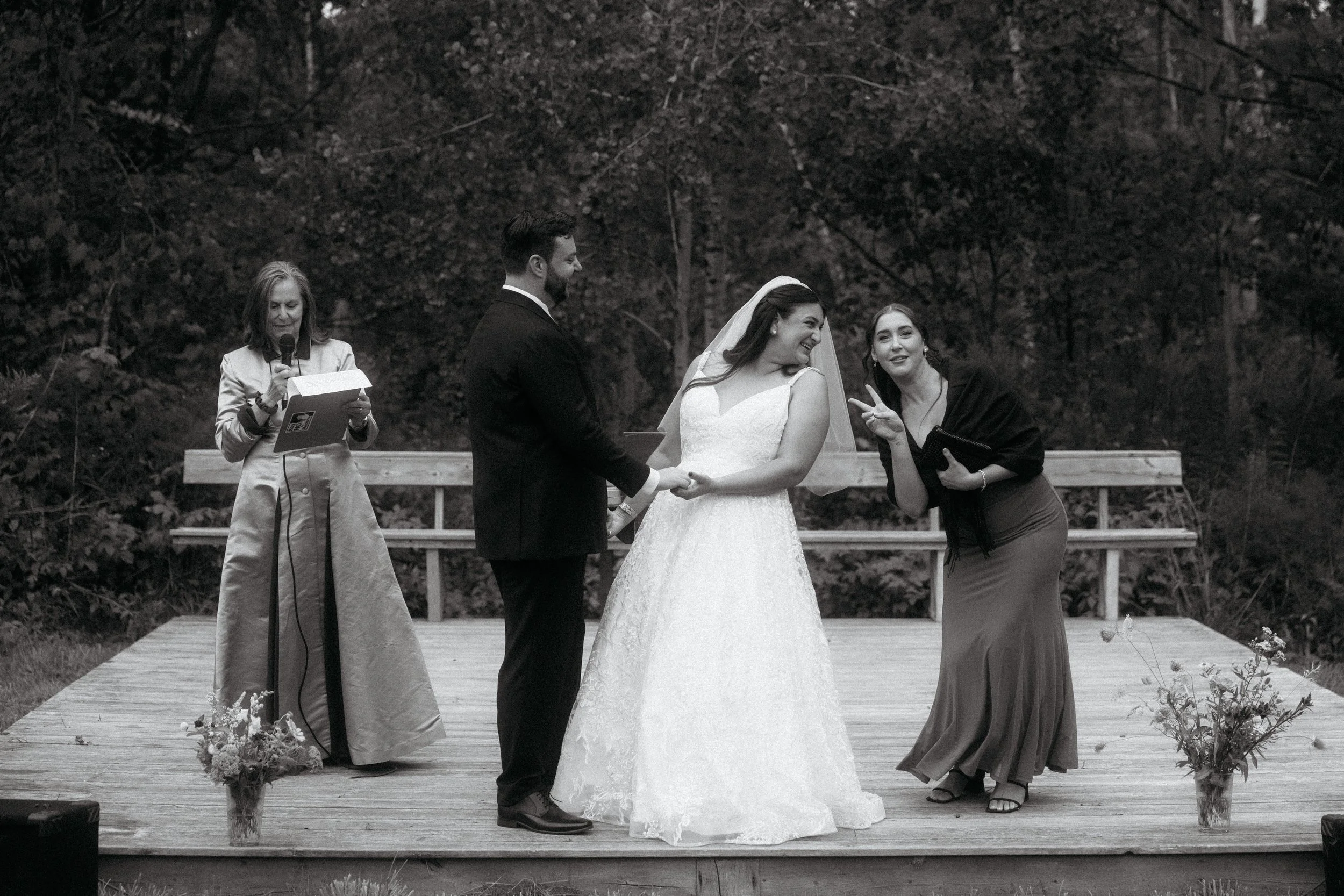 A black and white photo of a wedding ceremony outdoors with a bride and groom holding hands, a woman officiating, and a woman making a peace sign gesture.